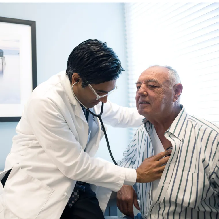Dr. Theuruvath is leaning in to listen to a senior male patient heartbeat with a stethoscope. 