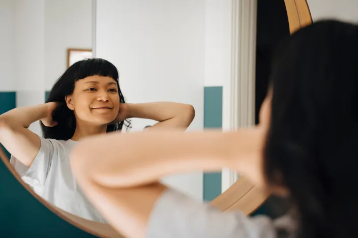 A young woman is pulling her hair back while looking at herself in the mirror.