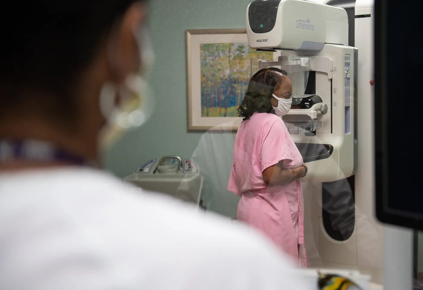 A Novant Health team member is standing behind a screen as a patient stands in a pink gown to have her mammogram screening.