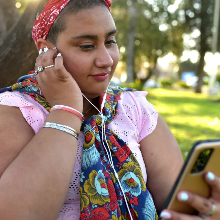 A teenager is sitting in a park adjusting the audio on her smart phone as she wears headphones. 