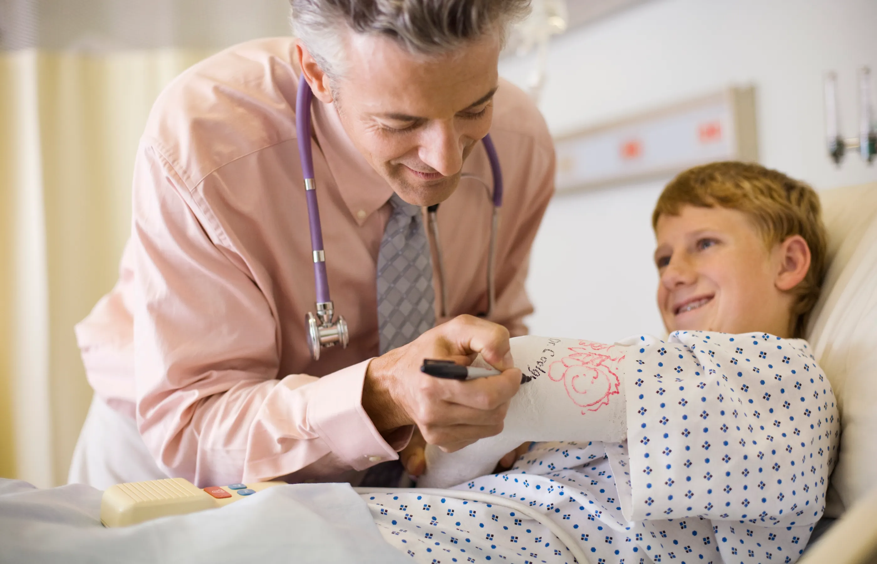 Pediatric orthopedic doctor signs an elementary age boy's broken arm as the boy lays in hospital bed. 
