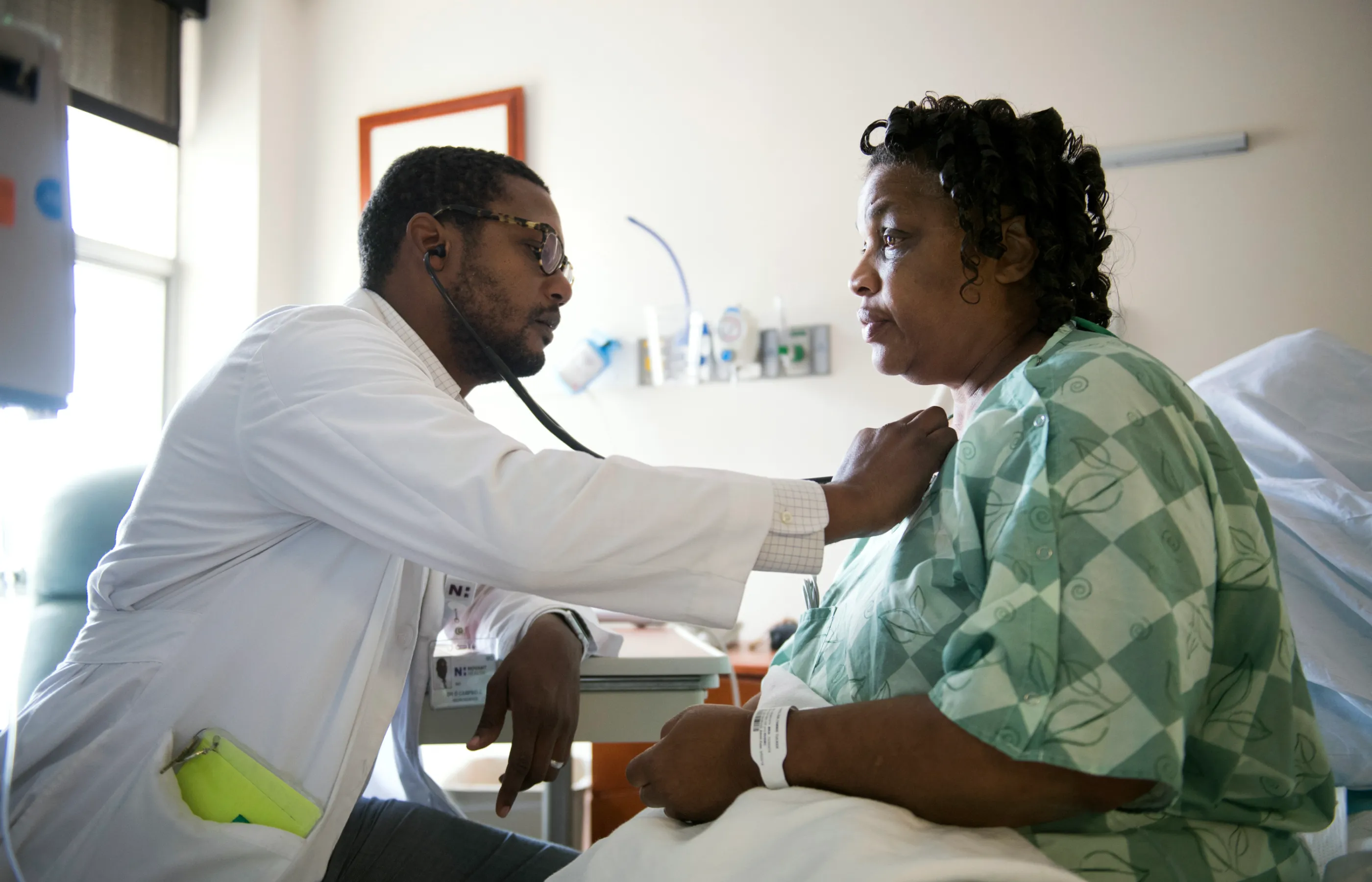 Dr. Campbell is sitting with a patient in their recovery room. With his stethoscope he listens to the patient's heart. 