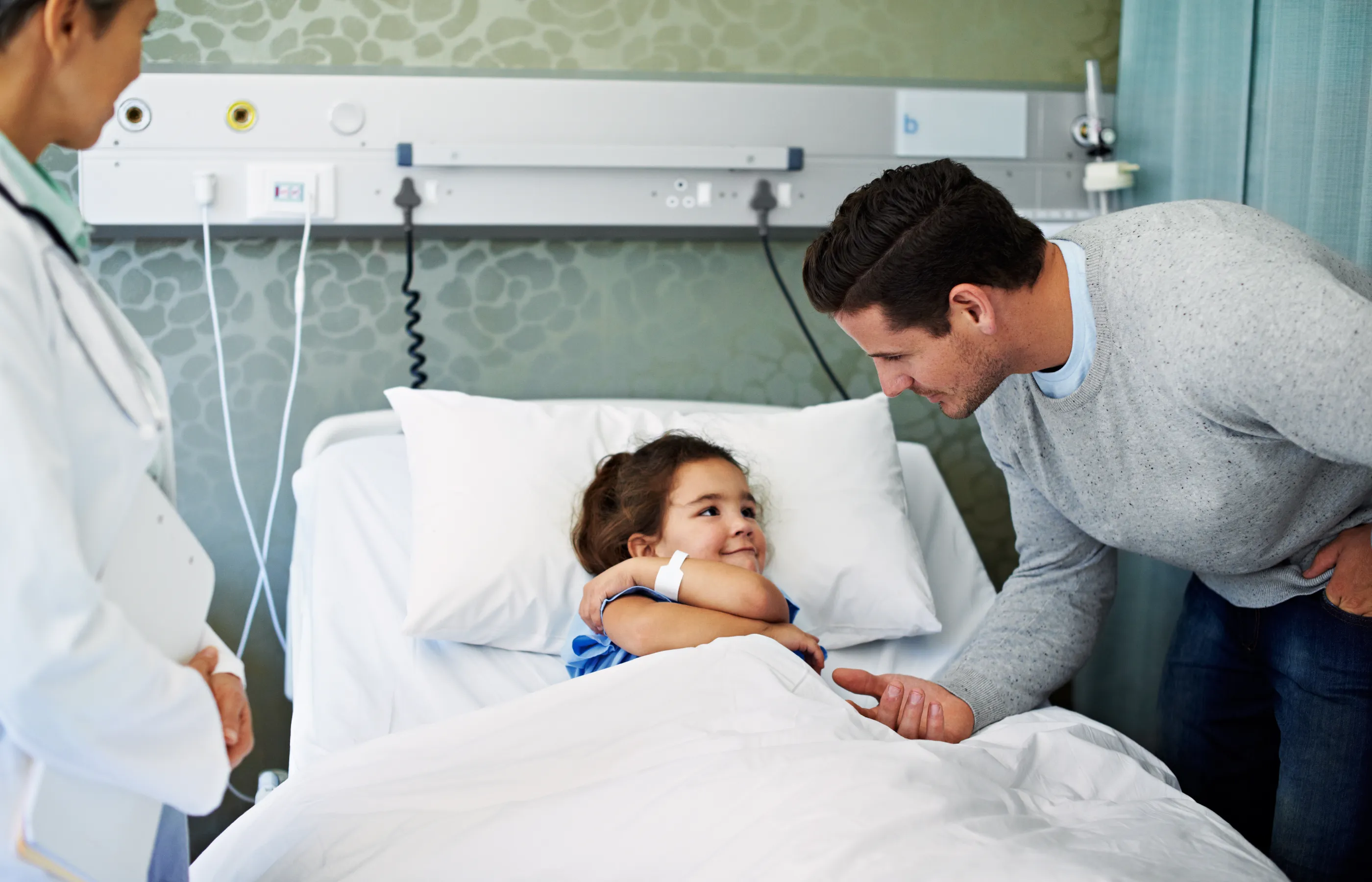 A father is comforter his daughter as she rests in a hospital bed. 