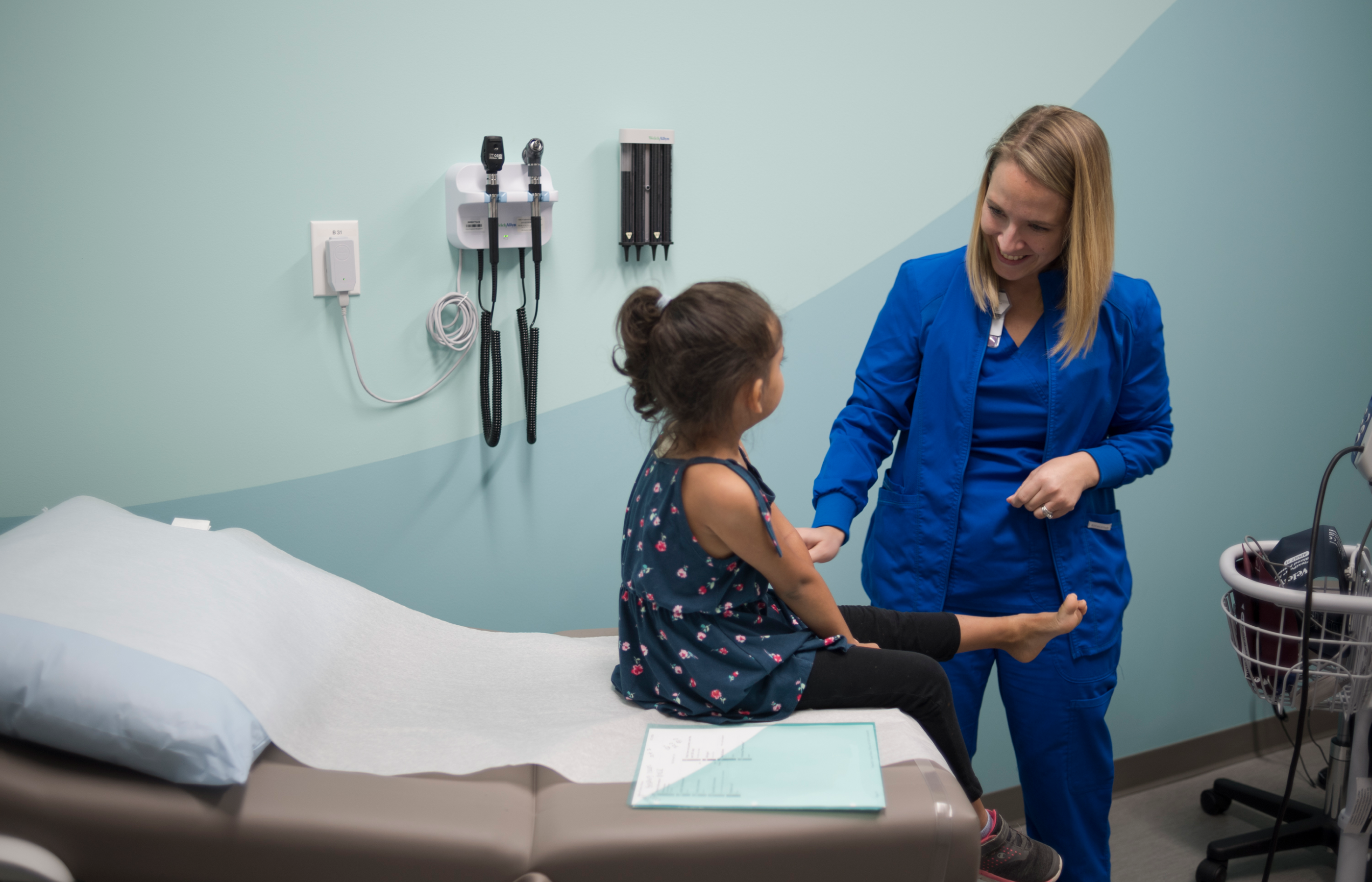 A young girl sits on an exam table while speaking with a nurse.