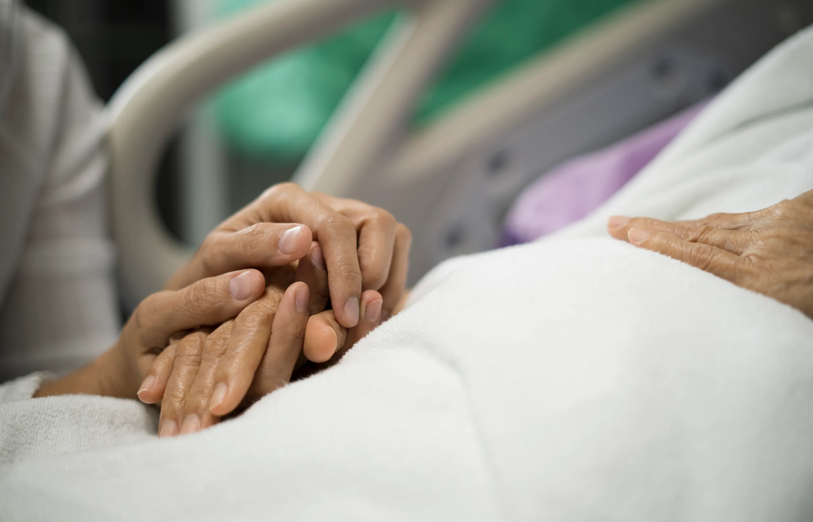 Patient in hospital bed with visitor bed side. Visitors' hands are covering the patients hands in support. 