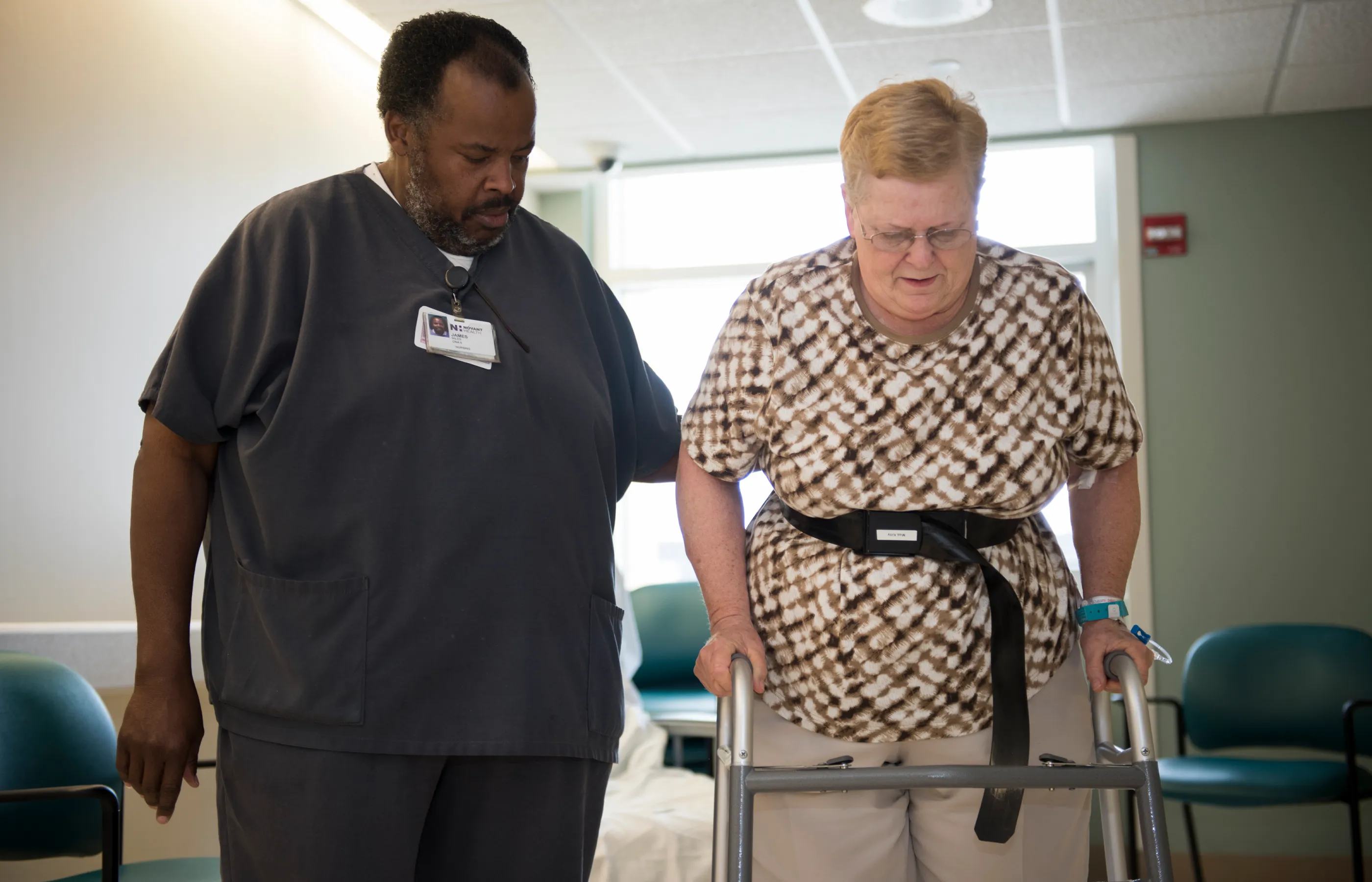 A male nurse is assistant a patient as she walks with a walker