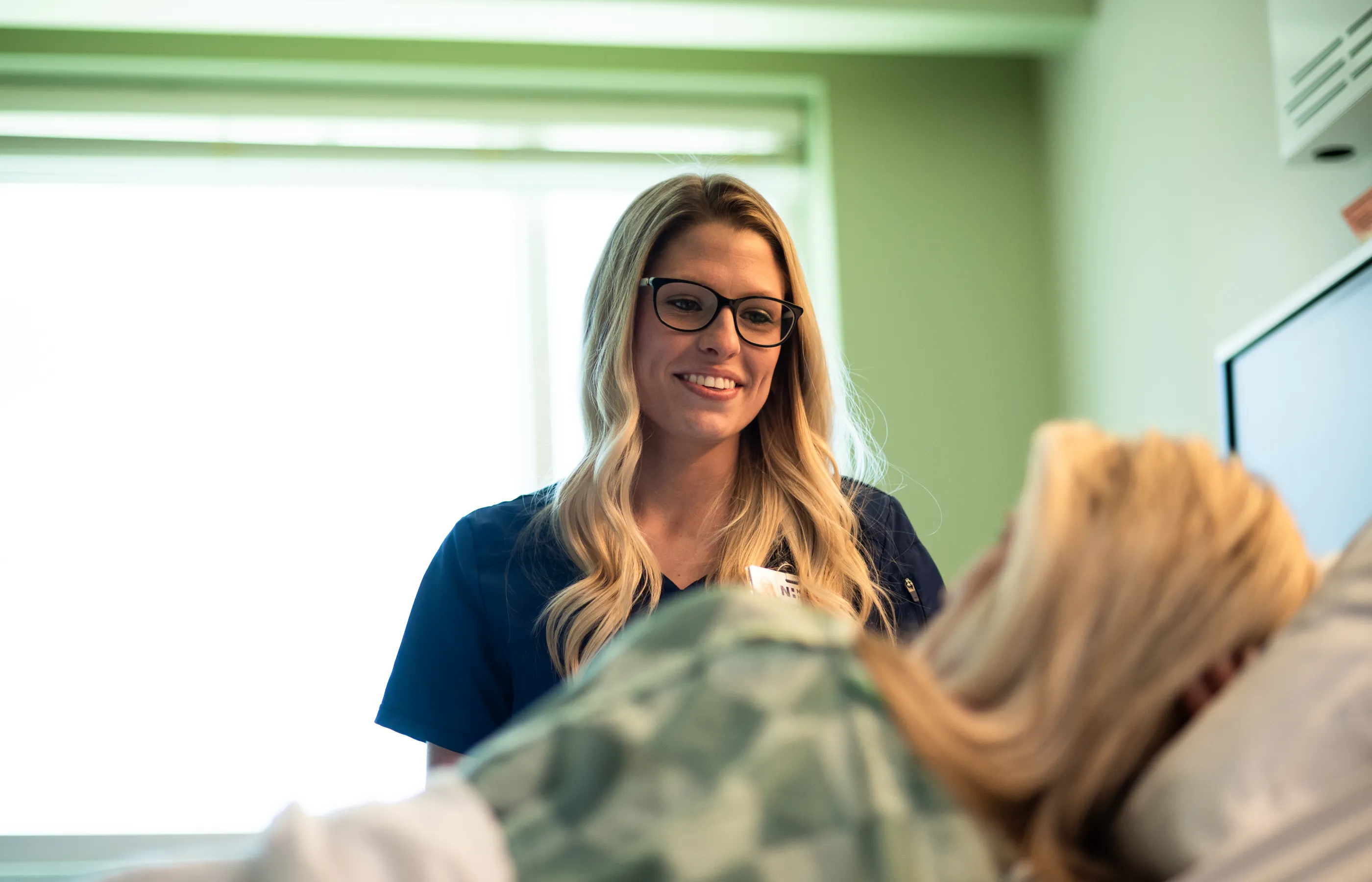 Novant Health nurse team member is standing in bedside in a patients recovery room. She smiles as she listens to the patient talk. 