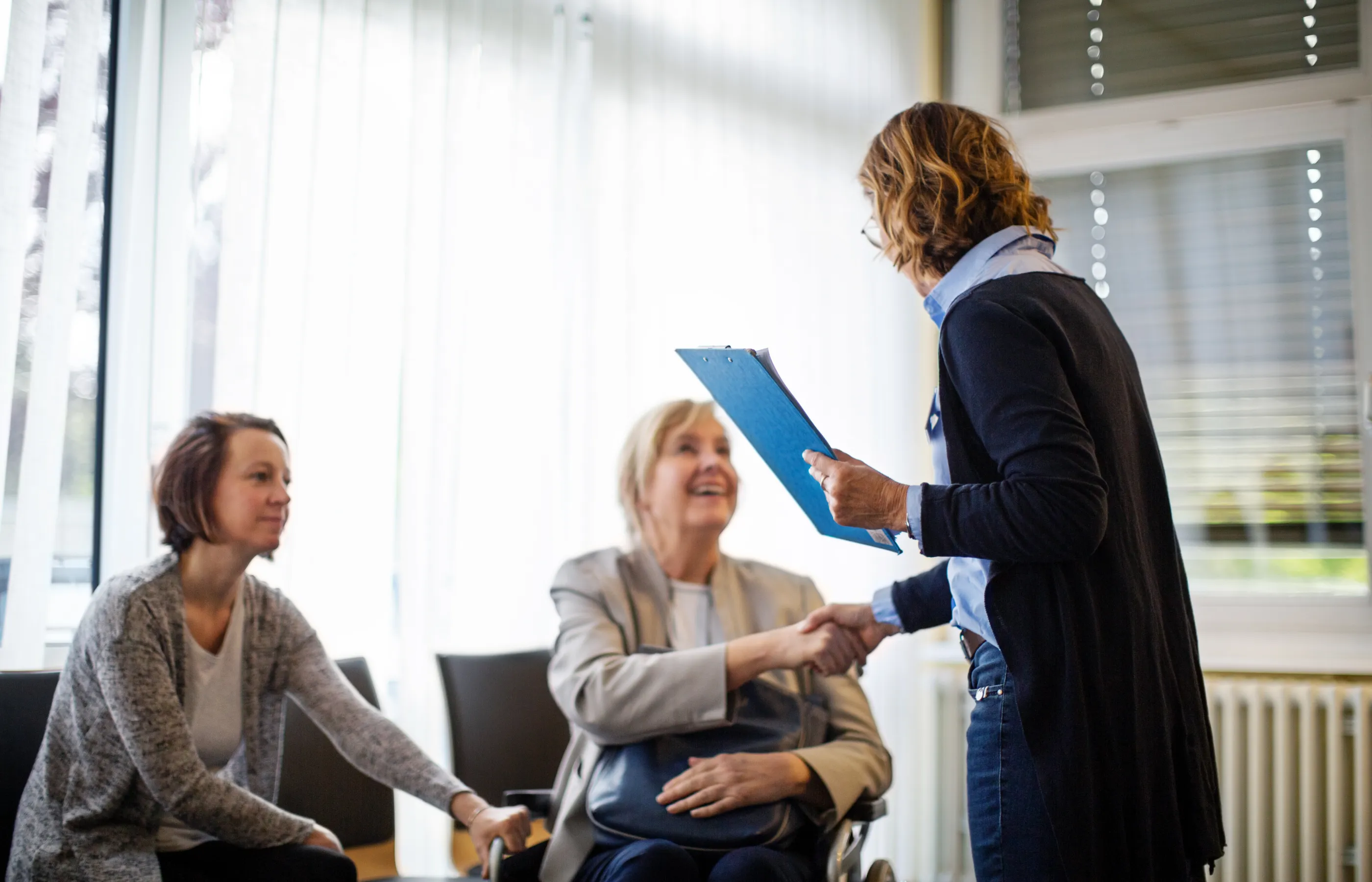 A senior woman and her adult daughter shake the hand of doctor in a waiting room. 