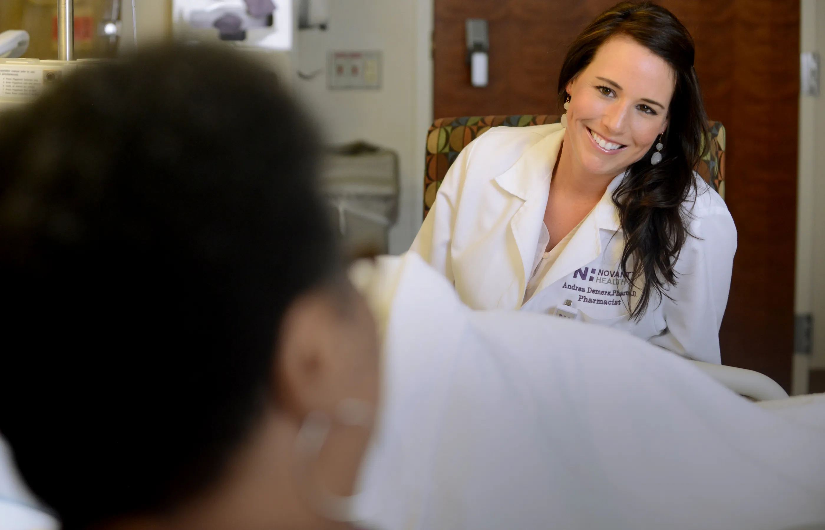 Novant Health pharmacist, Dr. Demera, is sitting at the bedside of a patient talking and smiling. 