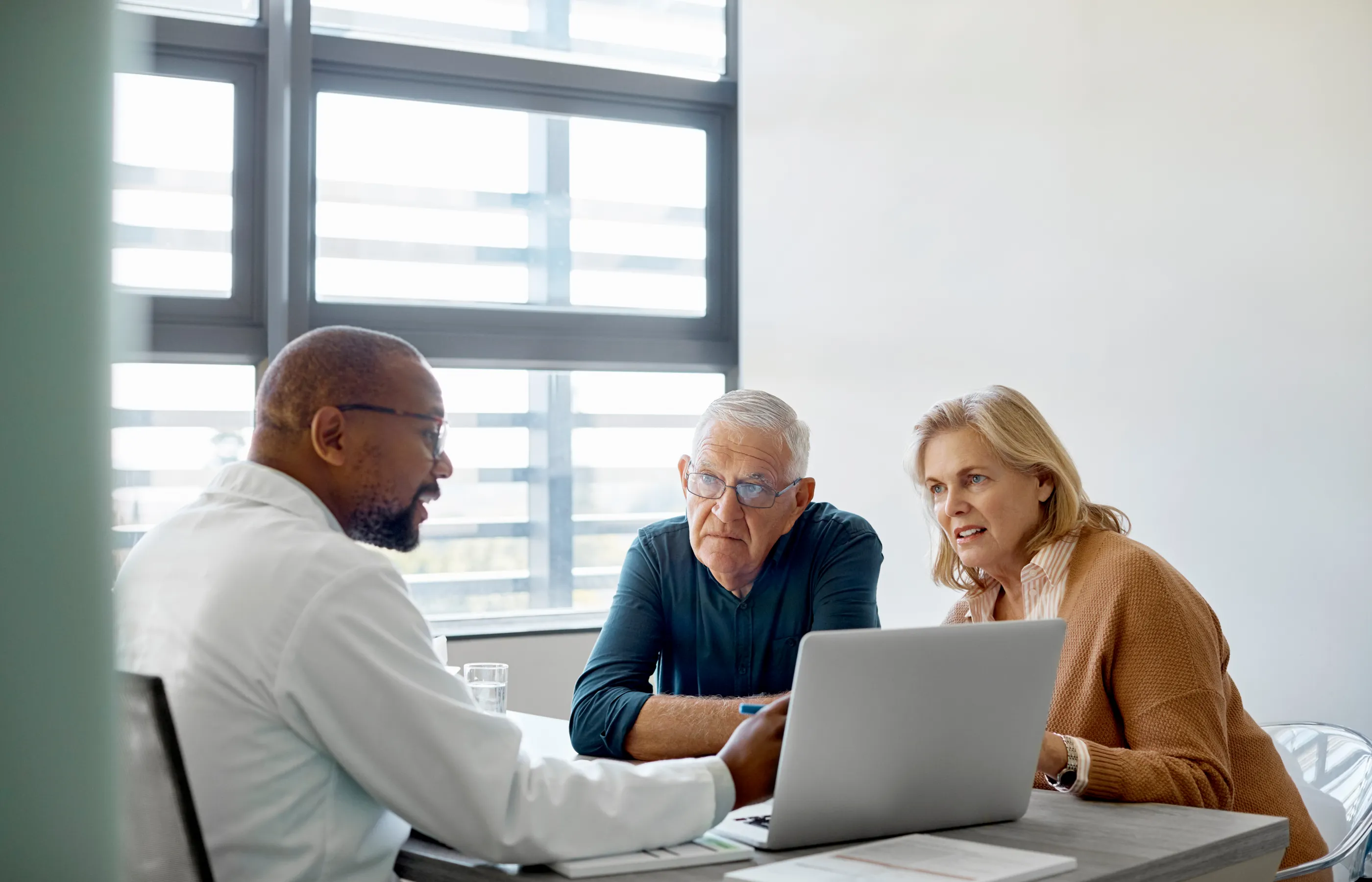 A senior couple are sitting across from a doctor as he shows them information on a laptop. 