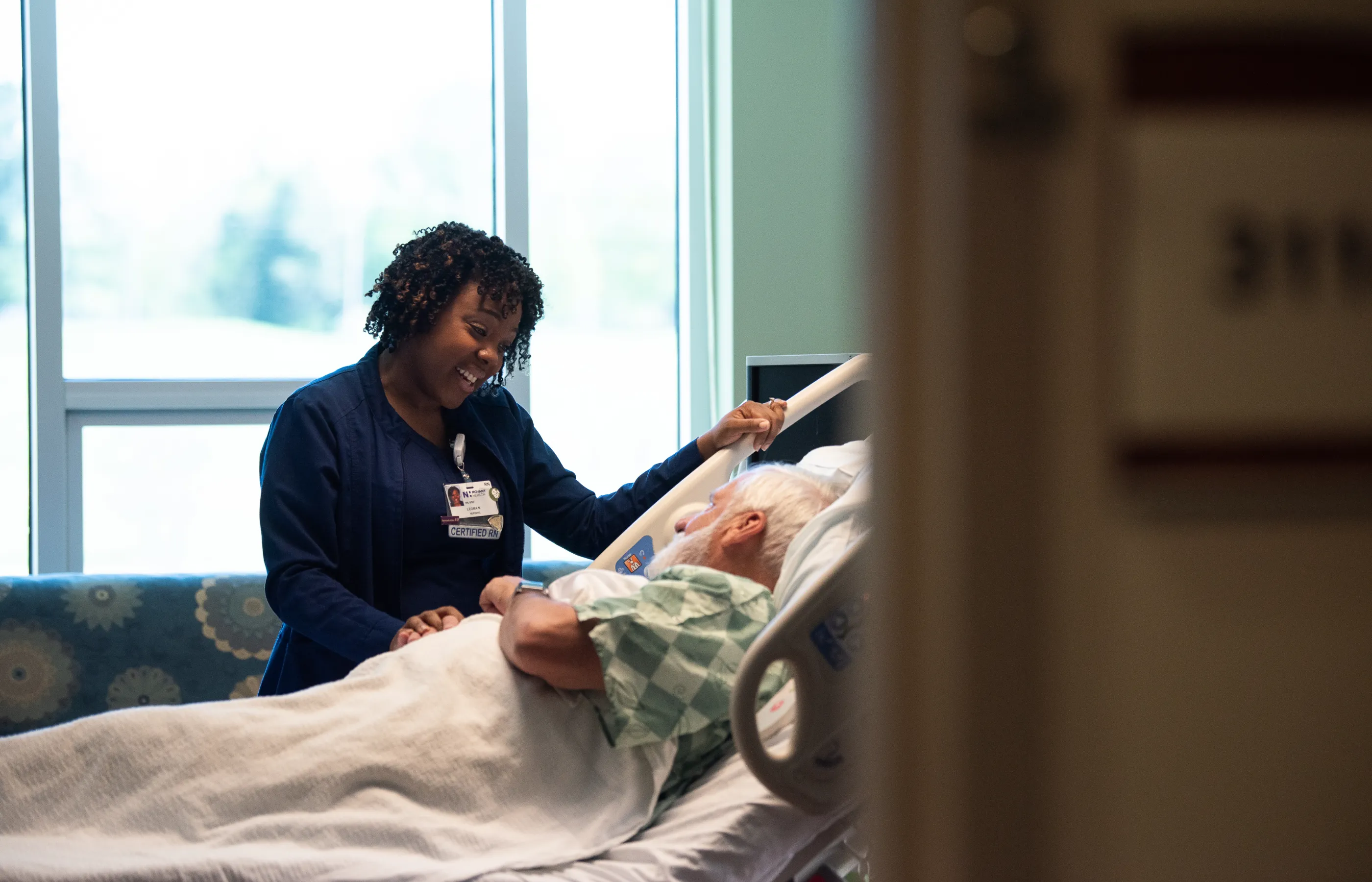A Novant Health nurse is standing at the bedside of a patient talking with the patient. 
