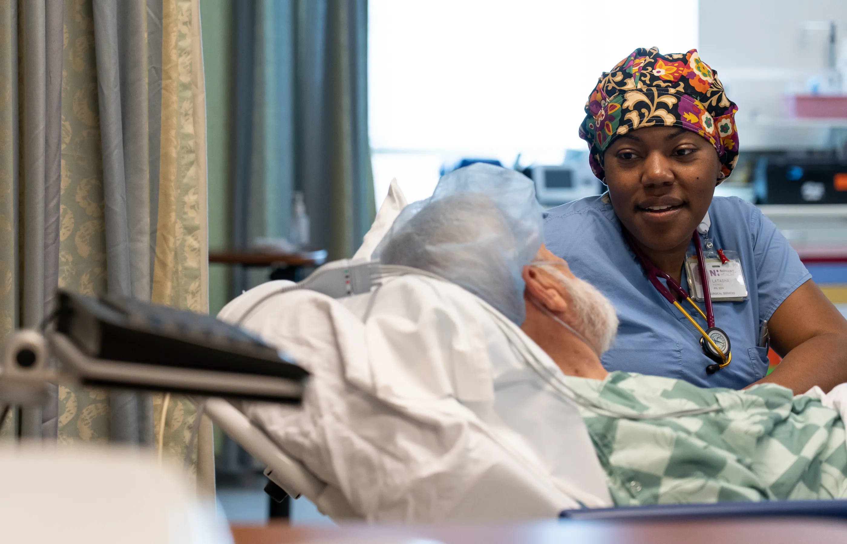 A Novant Health Registered Nurse is sitting at the bedside of a patient talking as they both prepare for surgery.