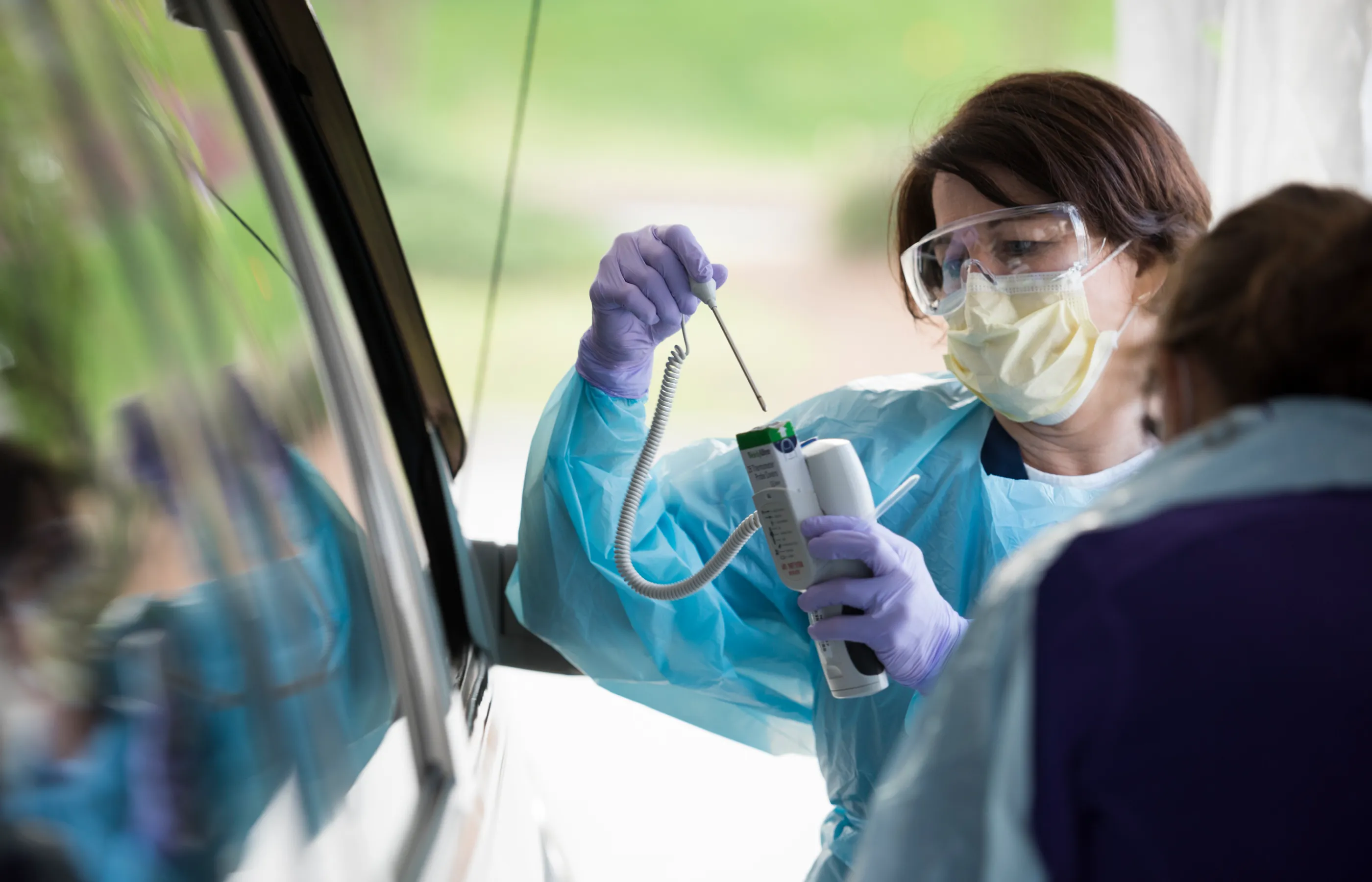 A team member wearing a mask, protective eyewear and gown looks at a thermometer next to a patient's car