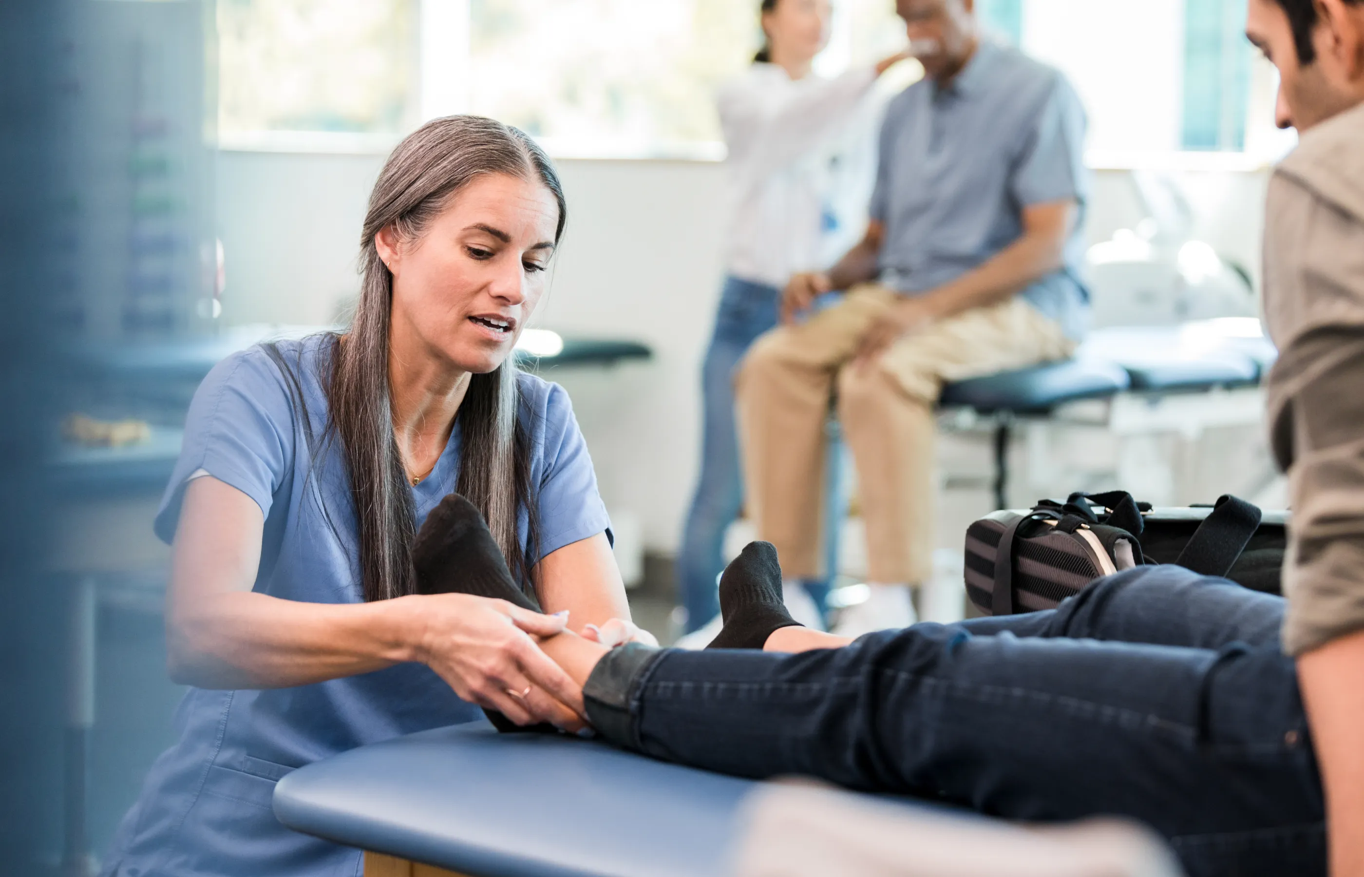 A team member squats next to a patient sitting on a table as she examines his ankle; another team member talks to another seated patient in the background