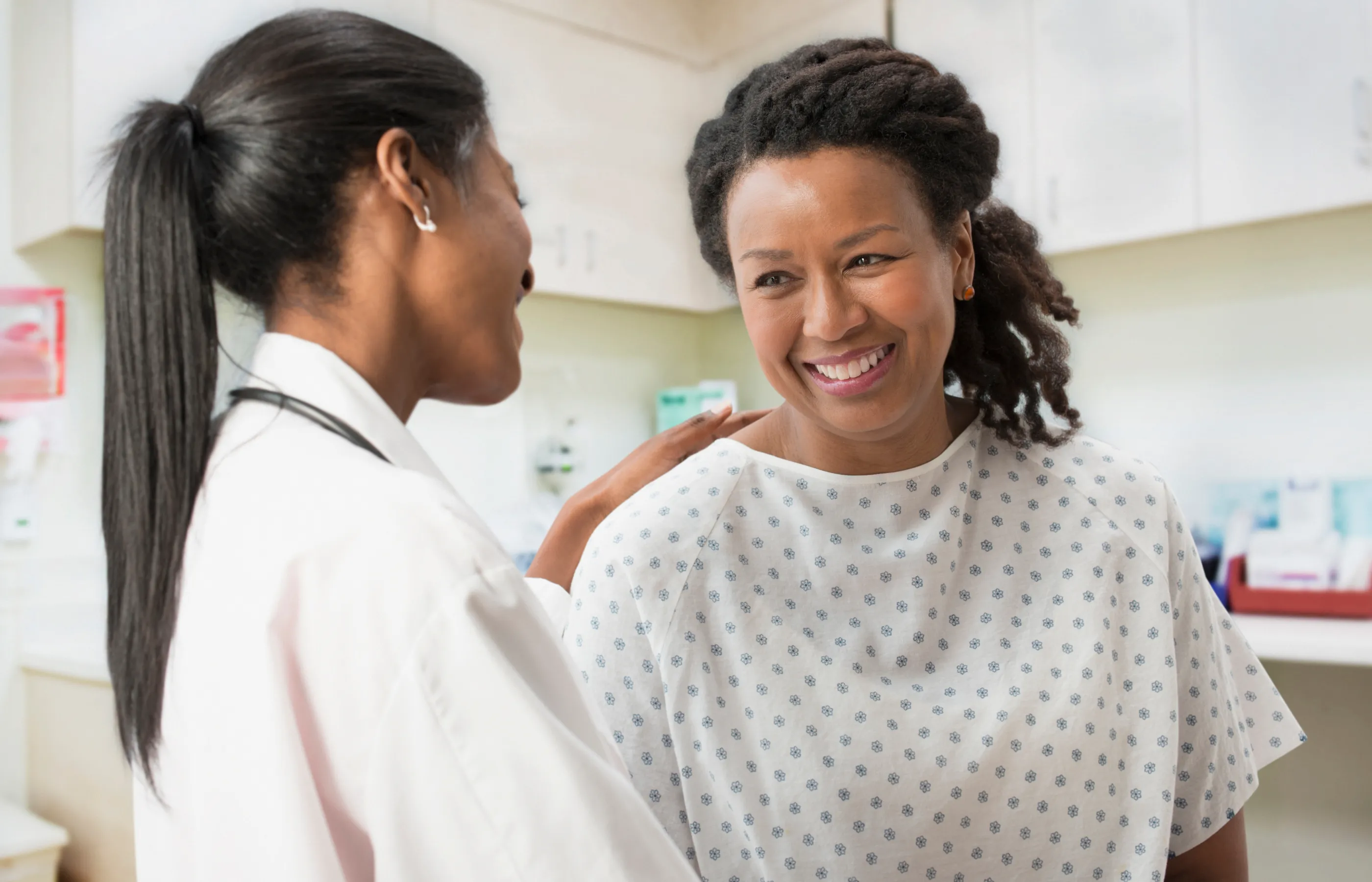 A woman, wearing a exam gown, is sitting and talking with her health care provider. 