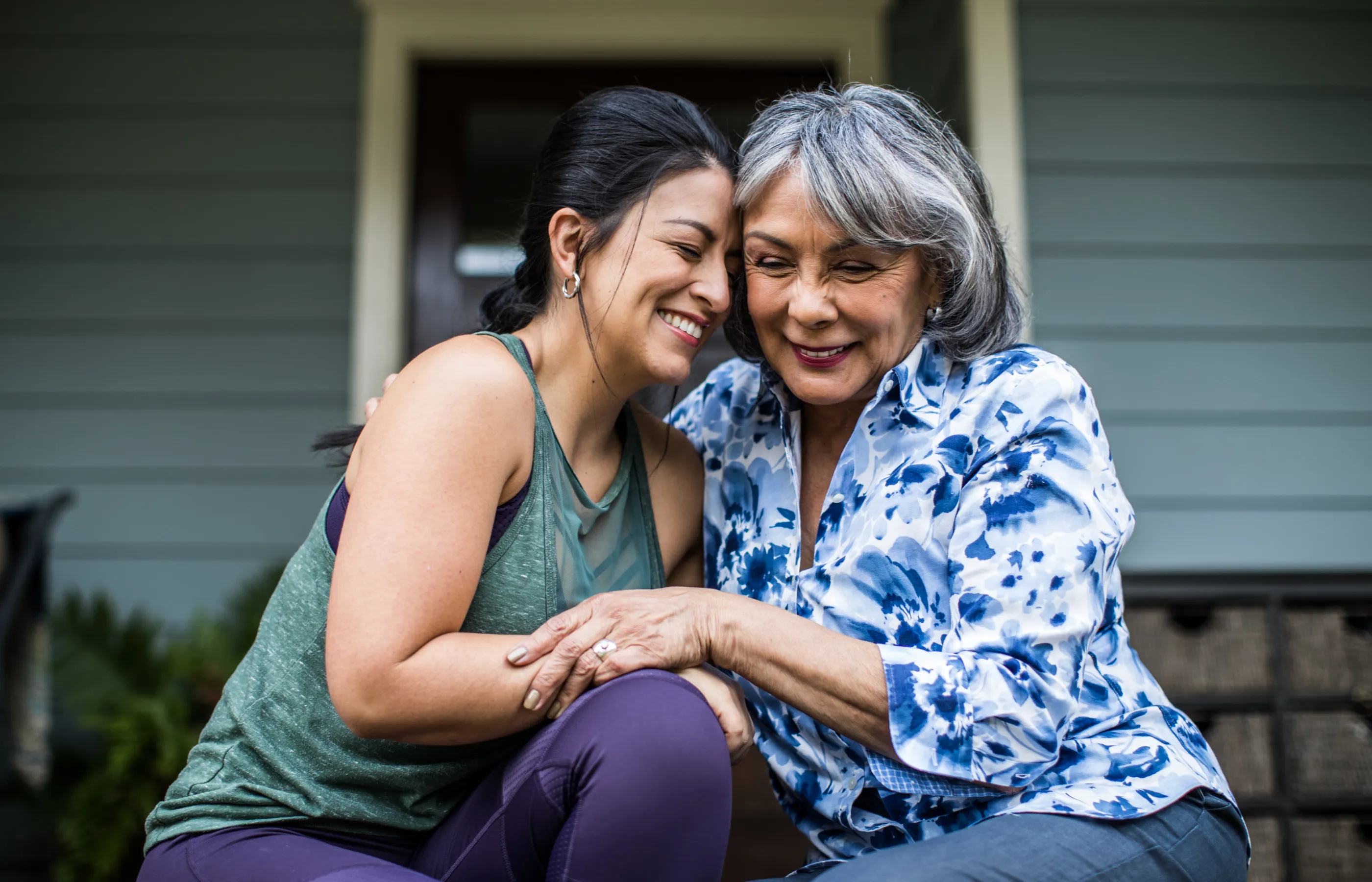 A mother, and her adult daughter, are sitting on their front porch smiling and hugging one another. 