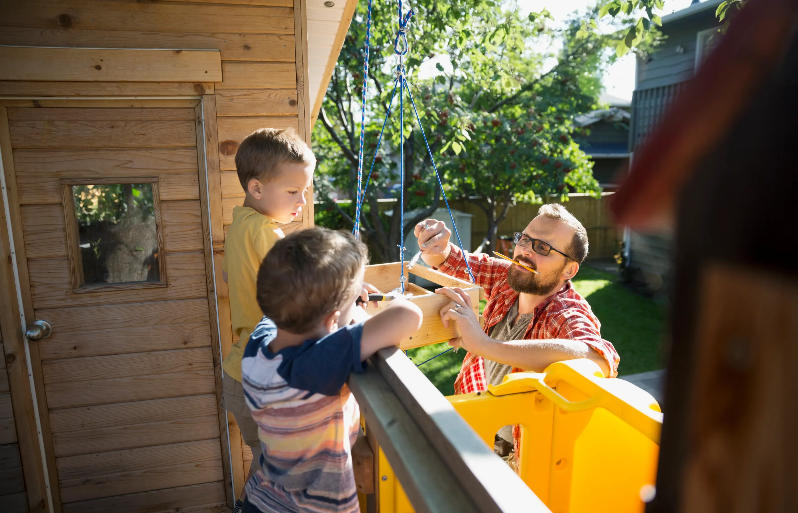 A dad is standing on a ladder with a pencil in his mother and tool in his hand. He is assisting his two young boys as they add parts to their tree house. 