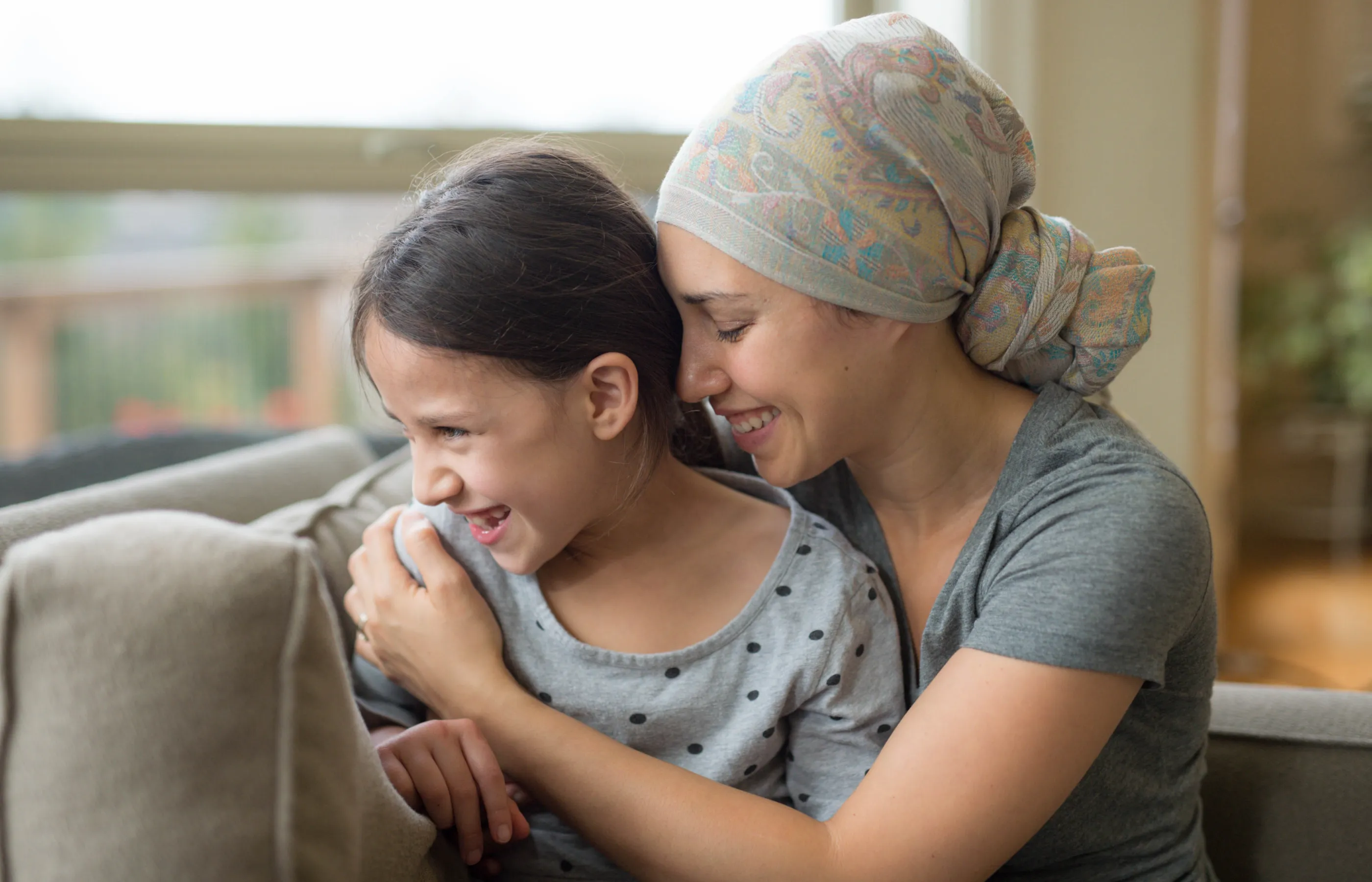 A mother, who is a cancer patient, is hugging her young daughter as they both look out the window of their home.