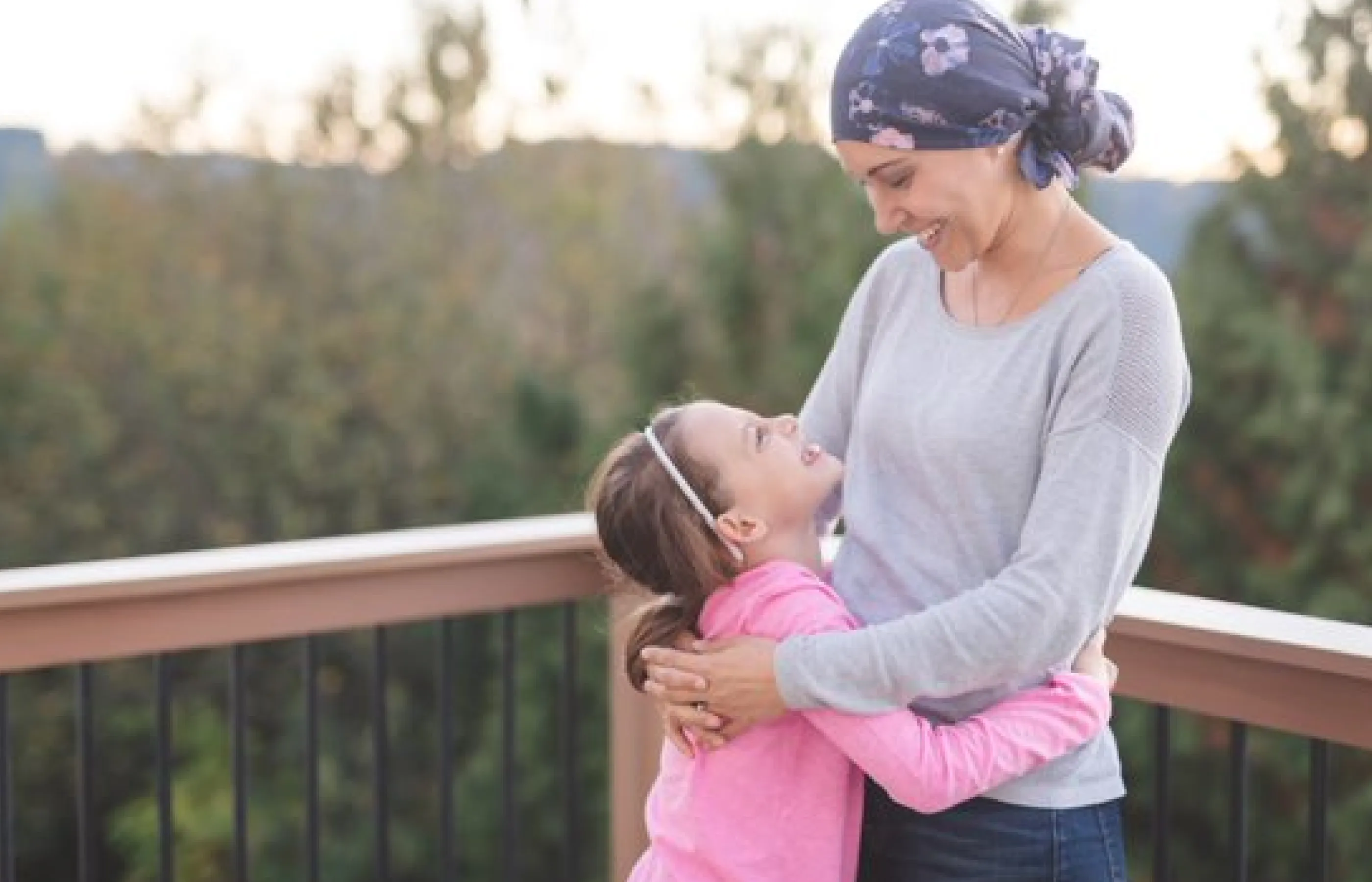 A young girl is looking up at her mother, who is wearing a headscarf due to cancer treatment. and smiling. Her mother is looking down at her and smiling back. 