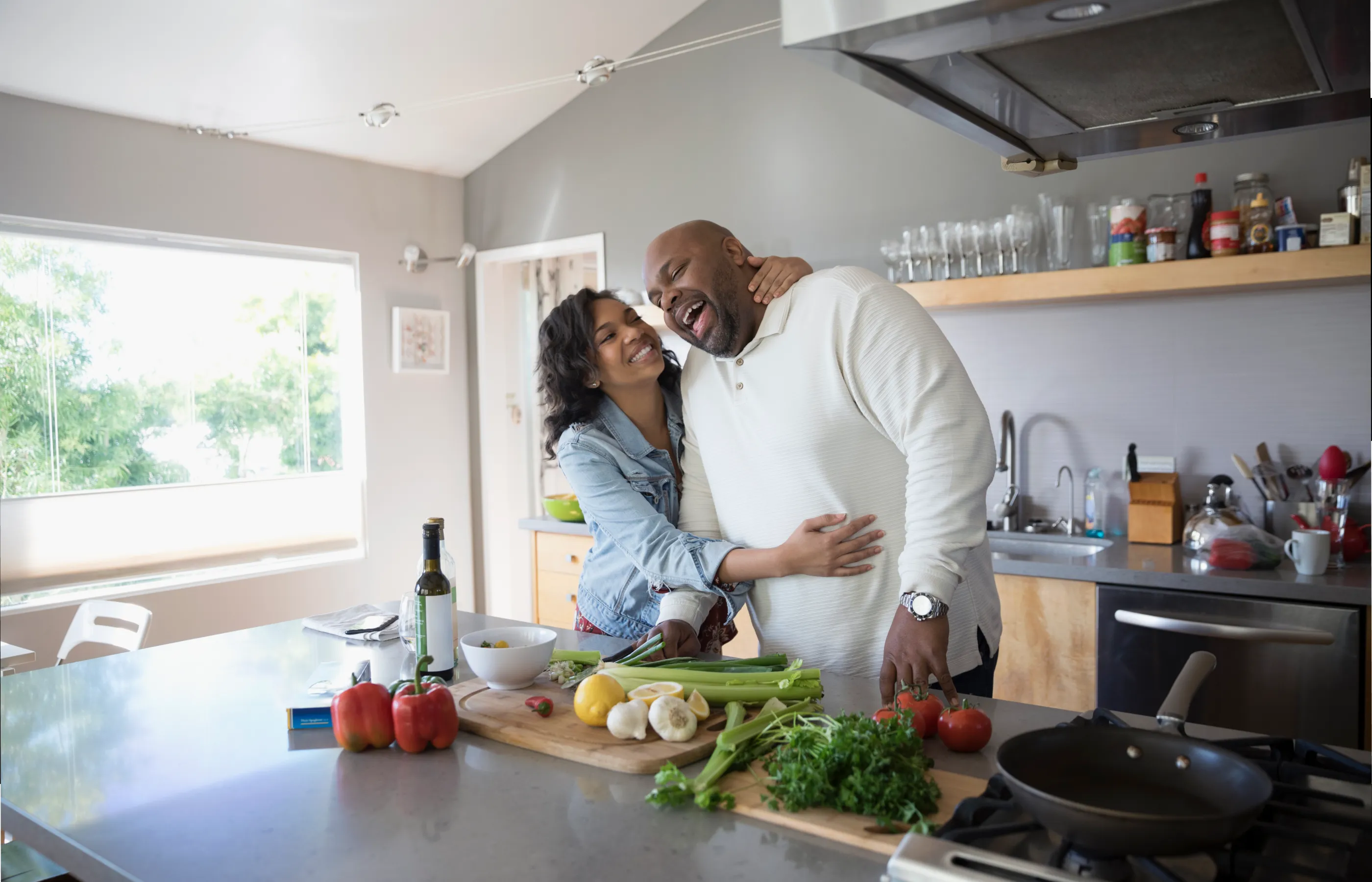 An African American couple is in the kitchen laughing together as they prepare a meal. 