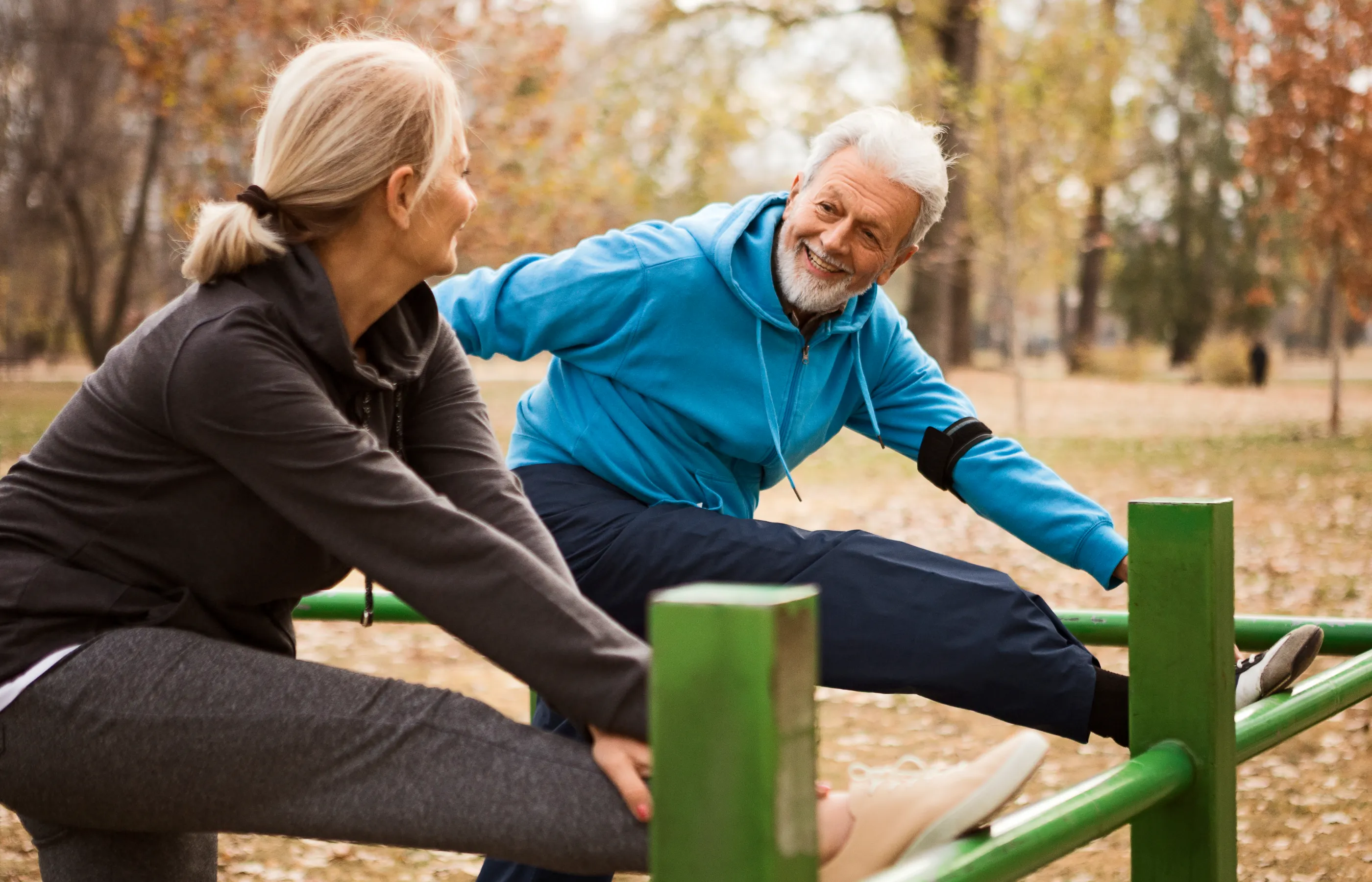 A senior couple is out at the park, in the fall. They both have one of their legs up on rails as they reach for their feet to stretch. 
