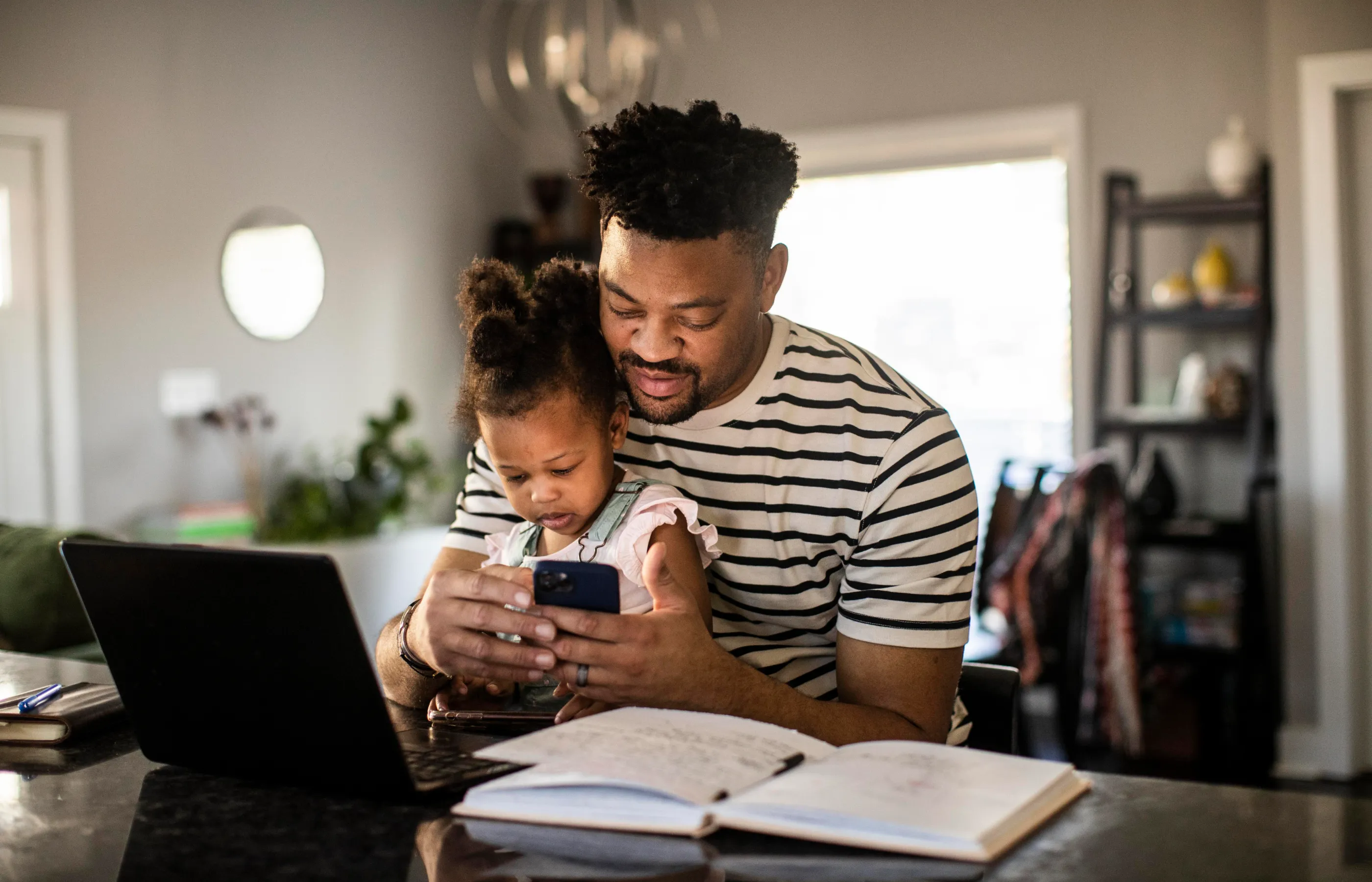 Father is sitting with his daughter at the counter. They are sitting in front of a laptop as he is looking for information on his phone.