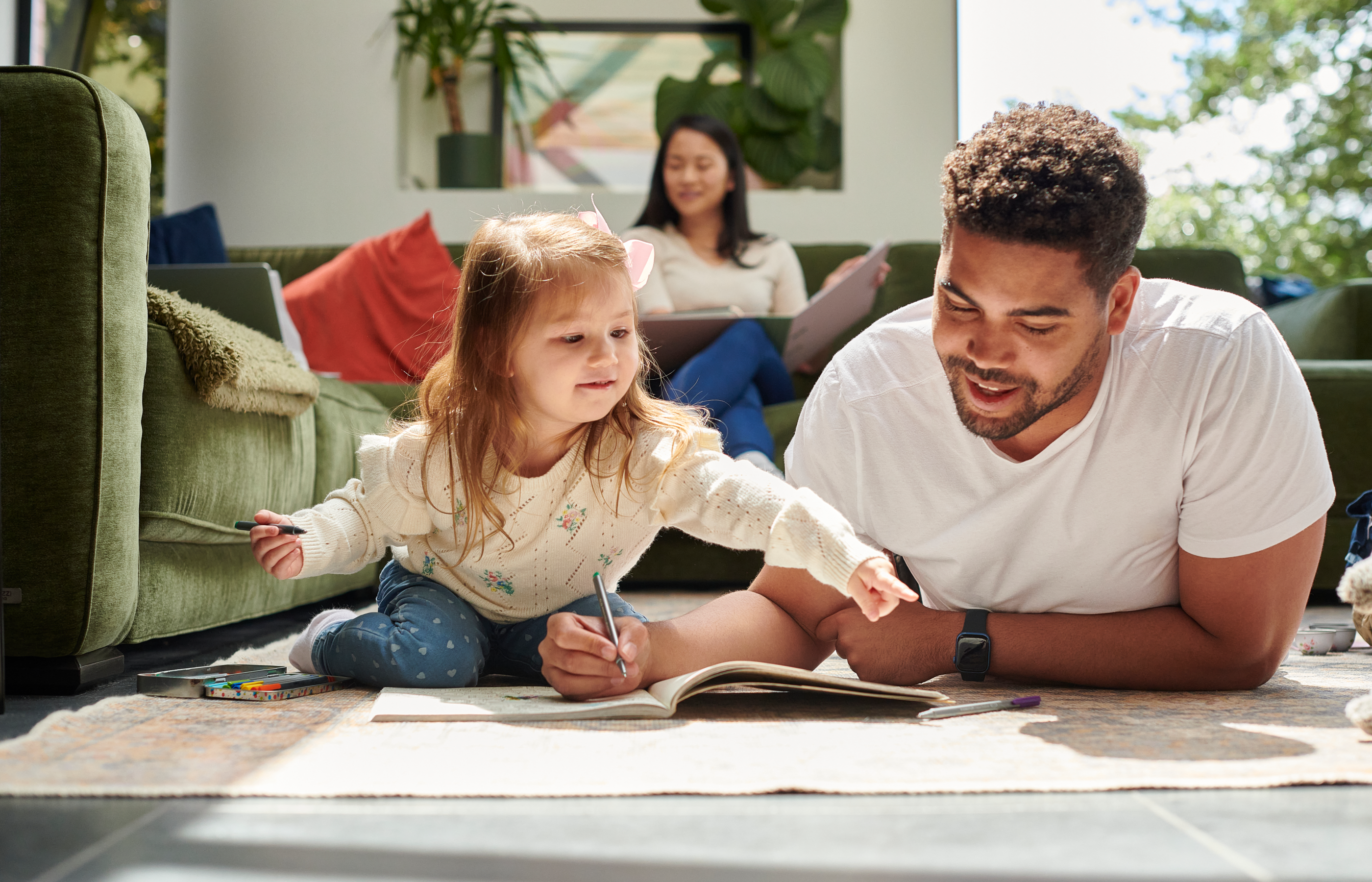 Father and daughter drawing together on the floor while mother watches from the couch in a sunlit, cozy room.