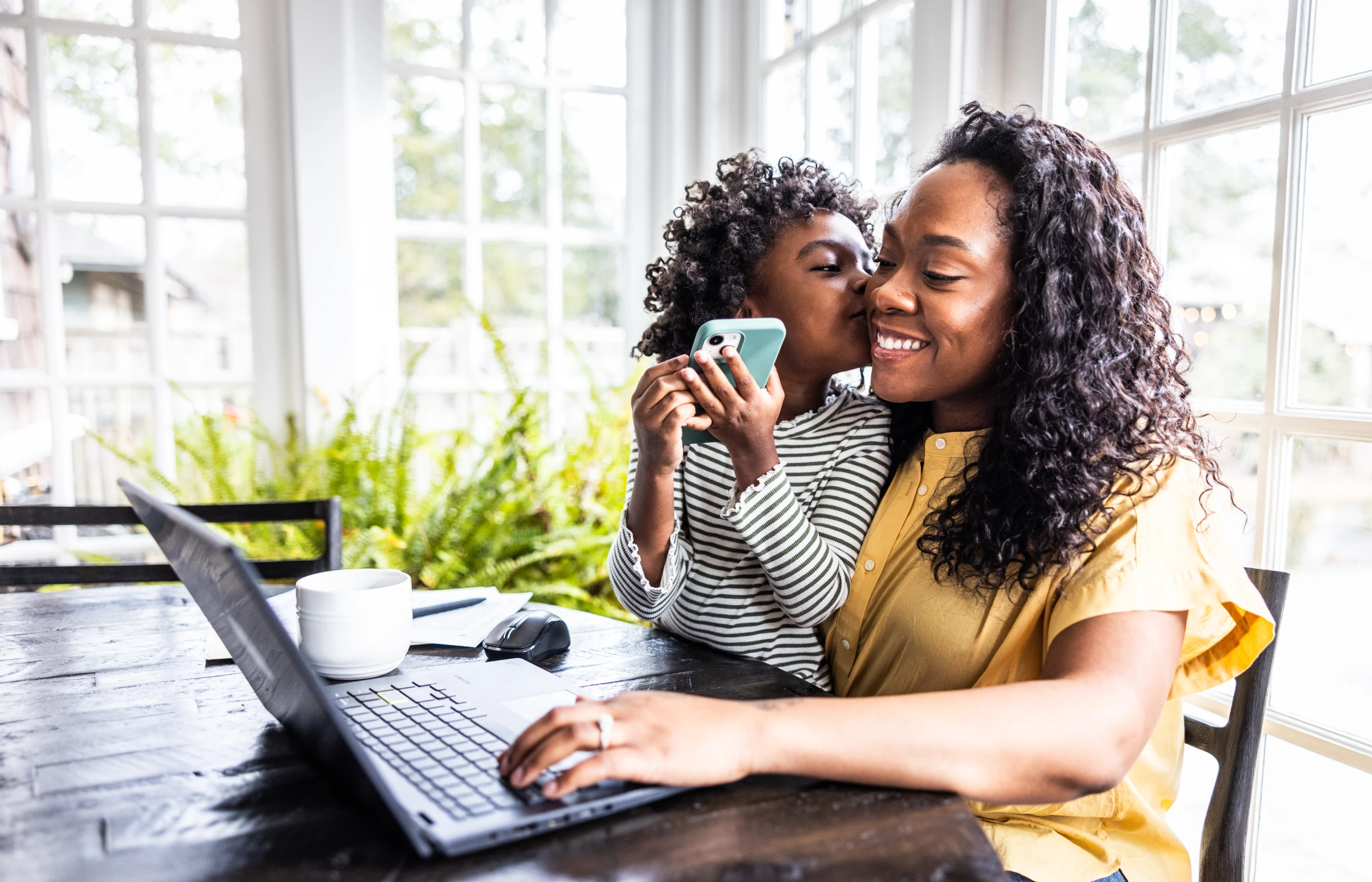 Female working on a laptop at home while a child stands nearby holding a smartphone.