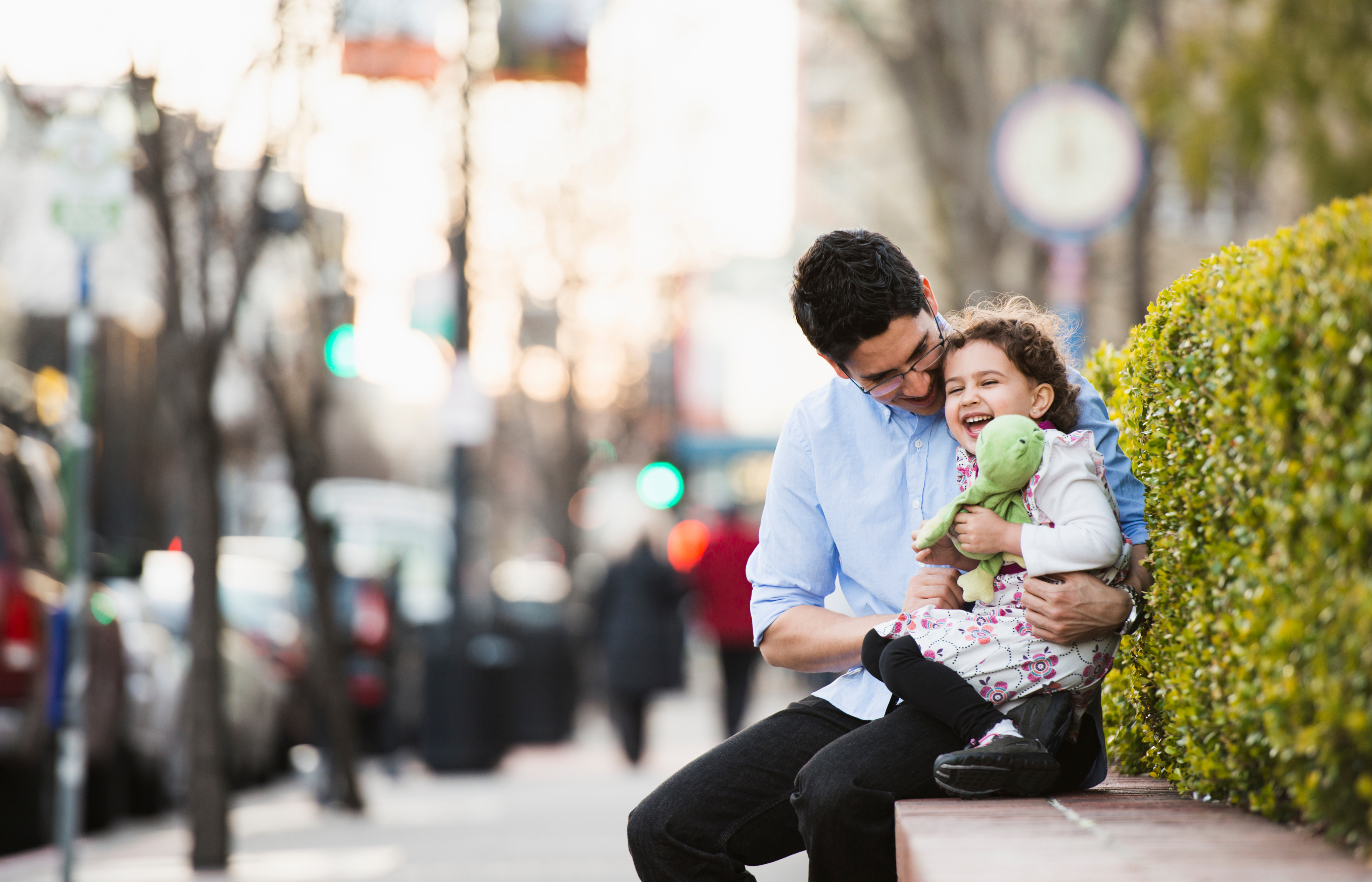 A father and his daughter sitting on a brick wall along a city sidewalk.