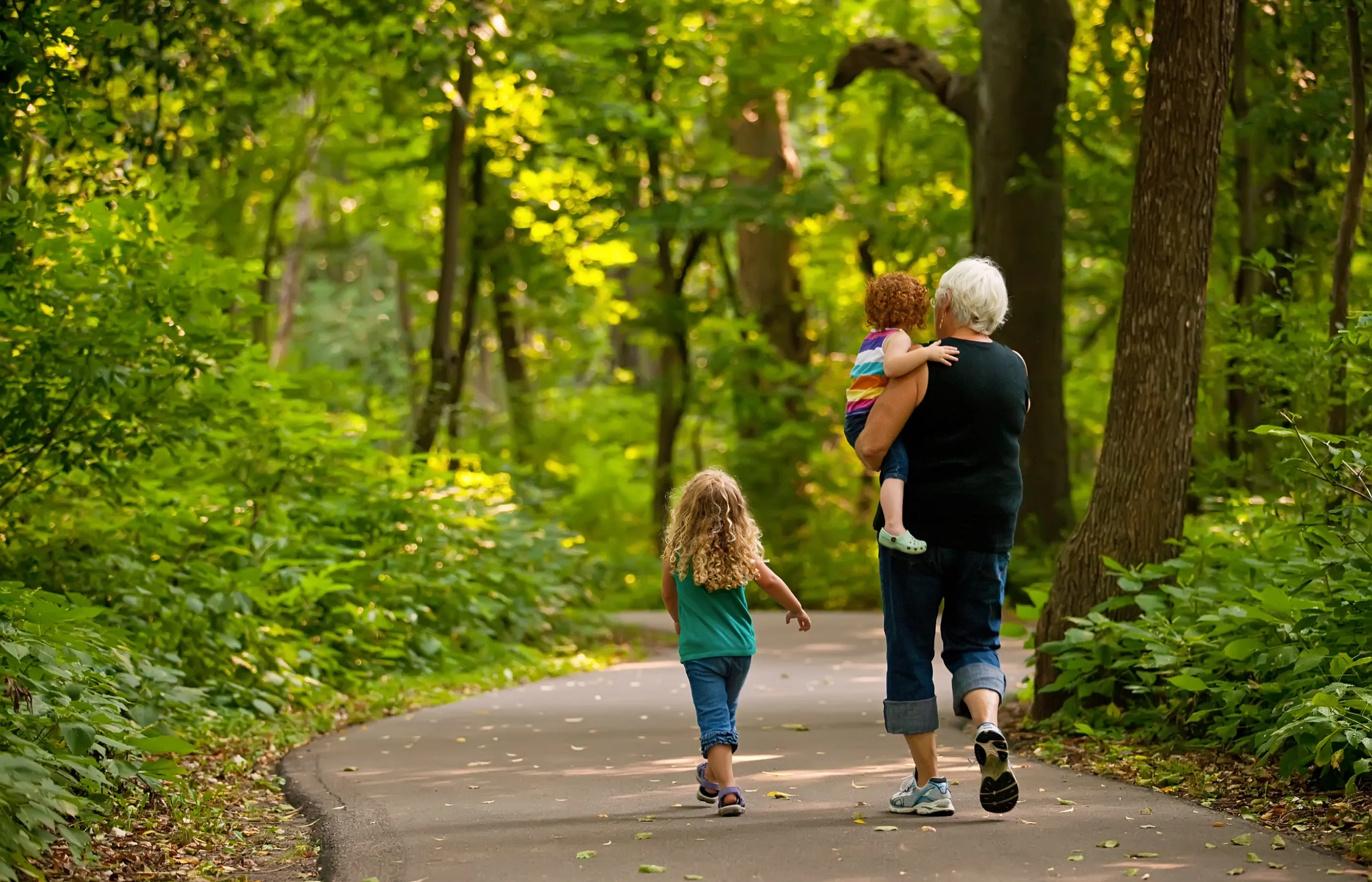 Grandmother is walking a greenway trail with her two grand children. One child is walking along side her as she carries the younger child.  