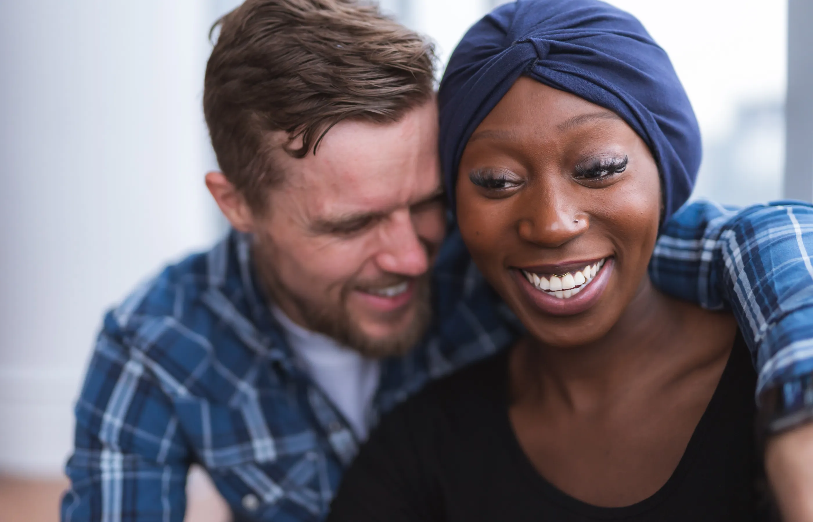 An interracial couple is sitting together, hugging, and smiling. The woman, who is a cancer patient, is wearing a turban on her head. 