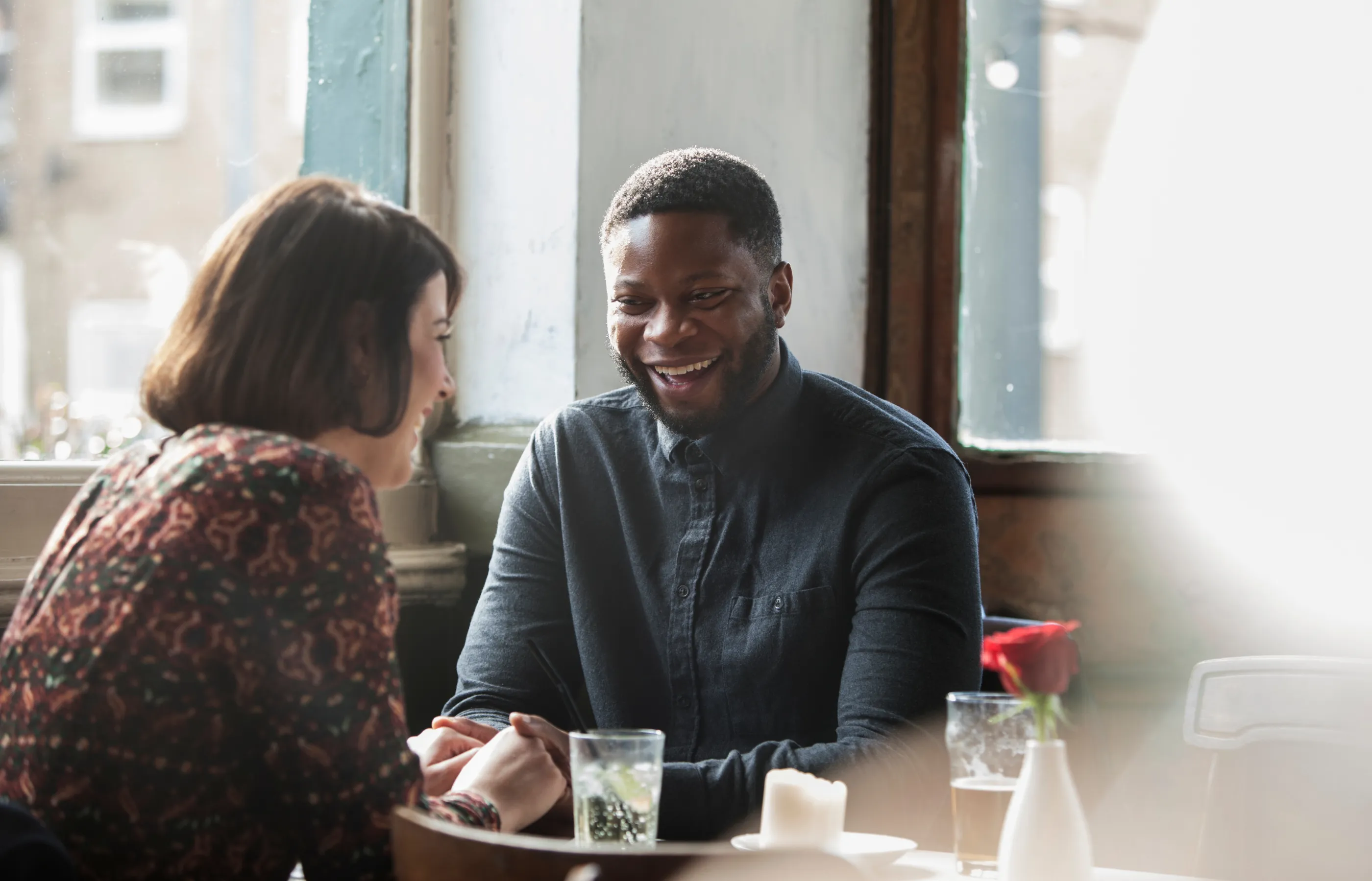 An interracial couple are sitting together at a restaurant, smiling, and holding hands. 