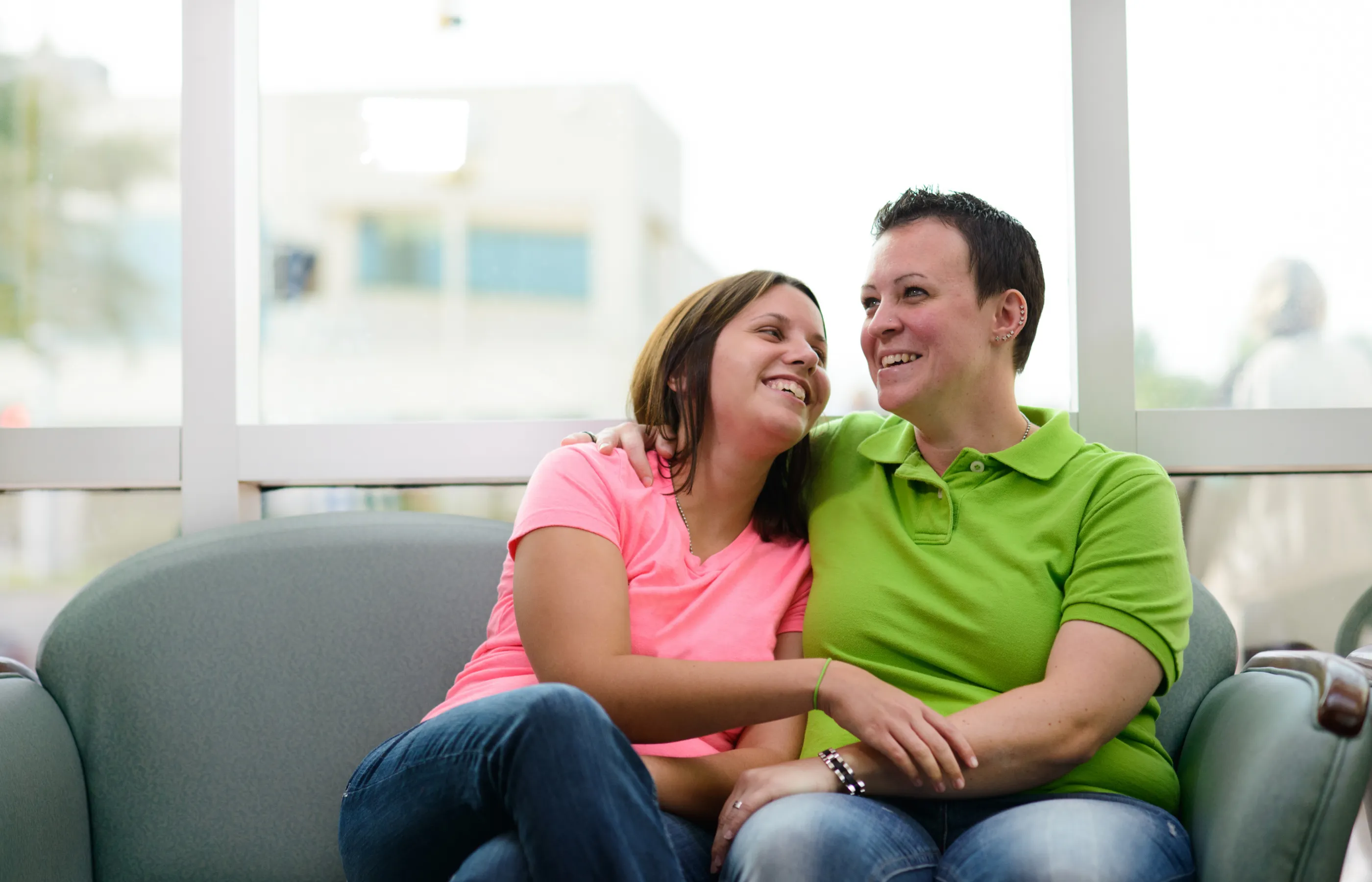 A same sex couple is sitting together on a couch, smiling, and embracing each other. 