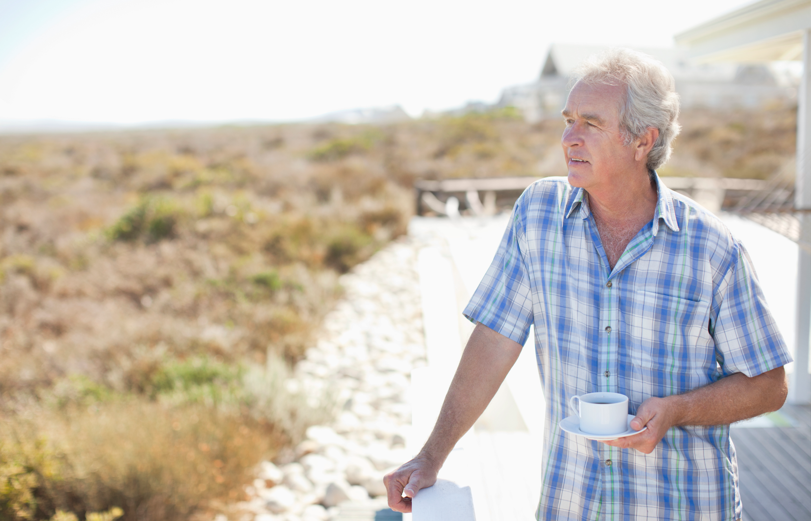 Person holding a white cup outdoors with dry grass and buildings in the background.