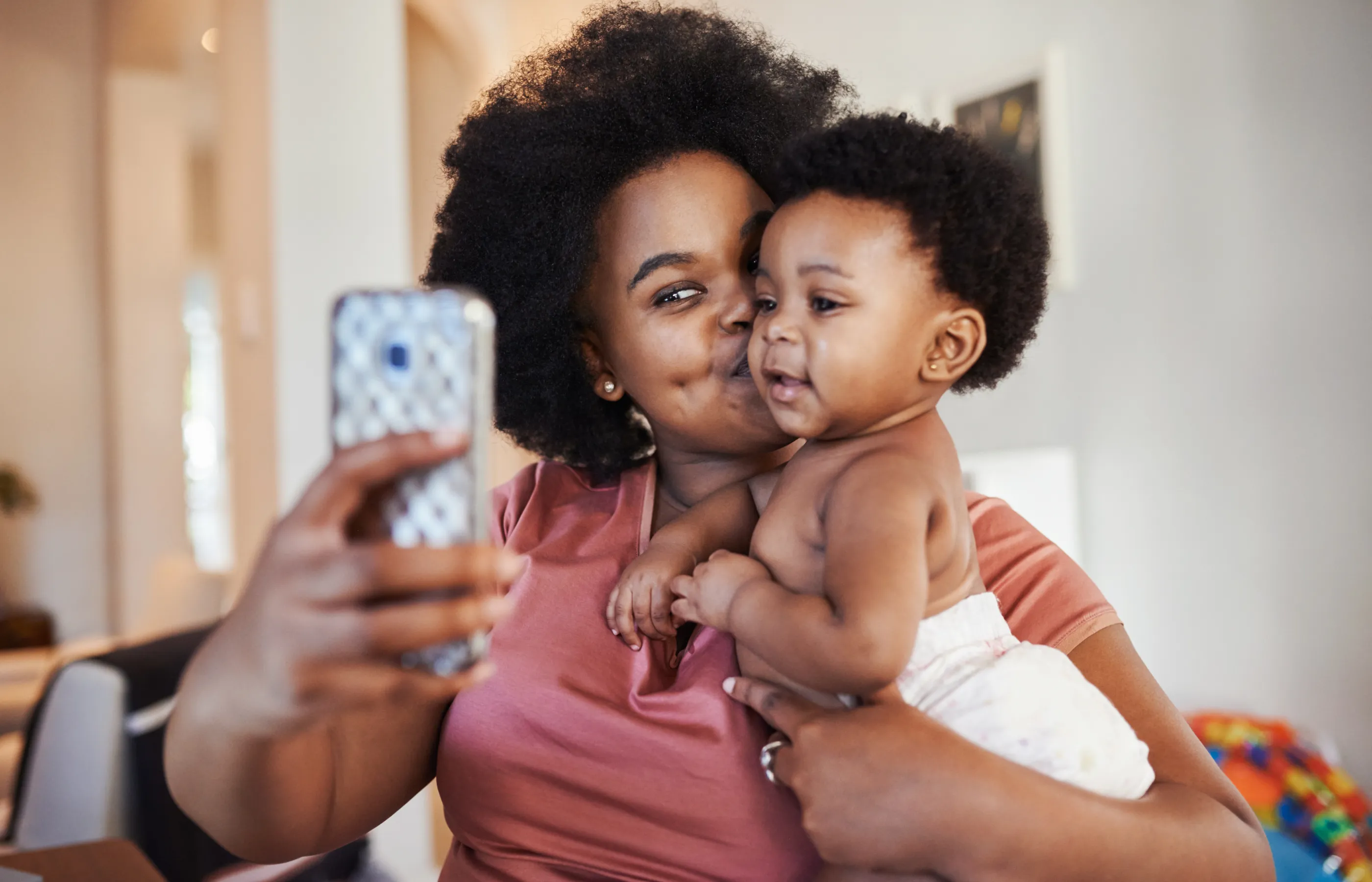 A mother is kissing and holding her baby while she takes a selfie. 