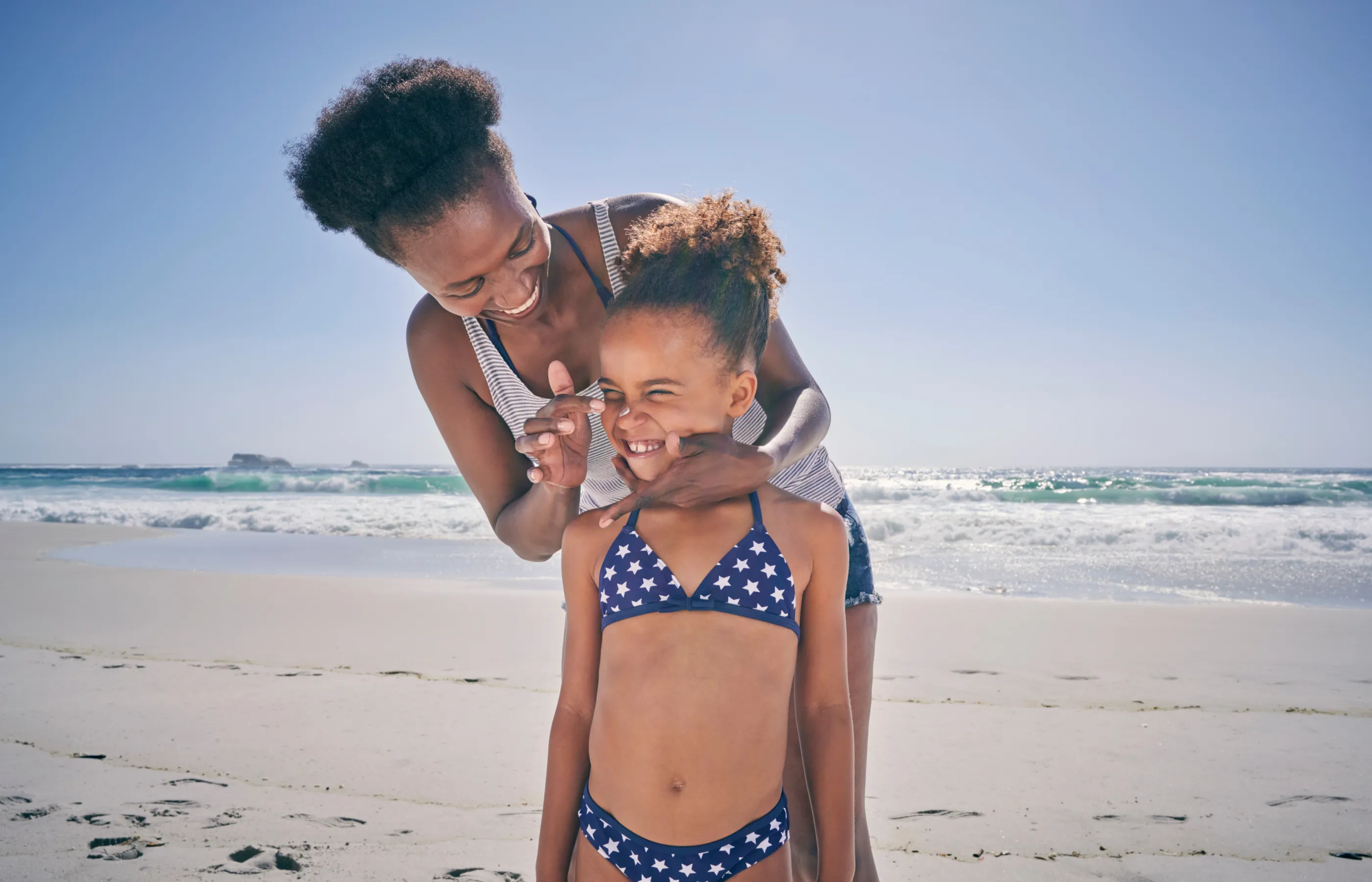 A mother and daughter are together at the beach. The mother has grabbed the face of her daughter to place sunscreen on her nose. 