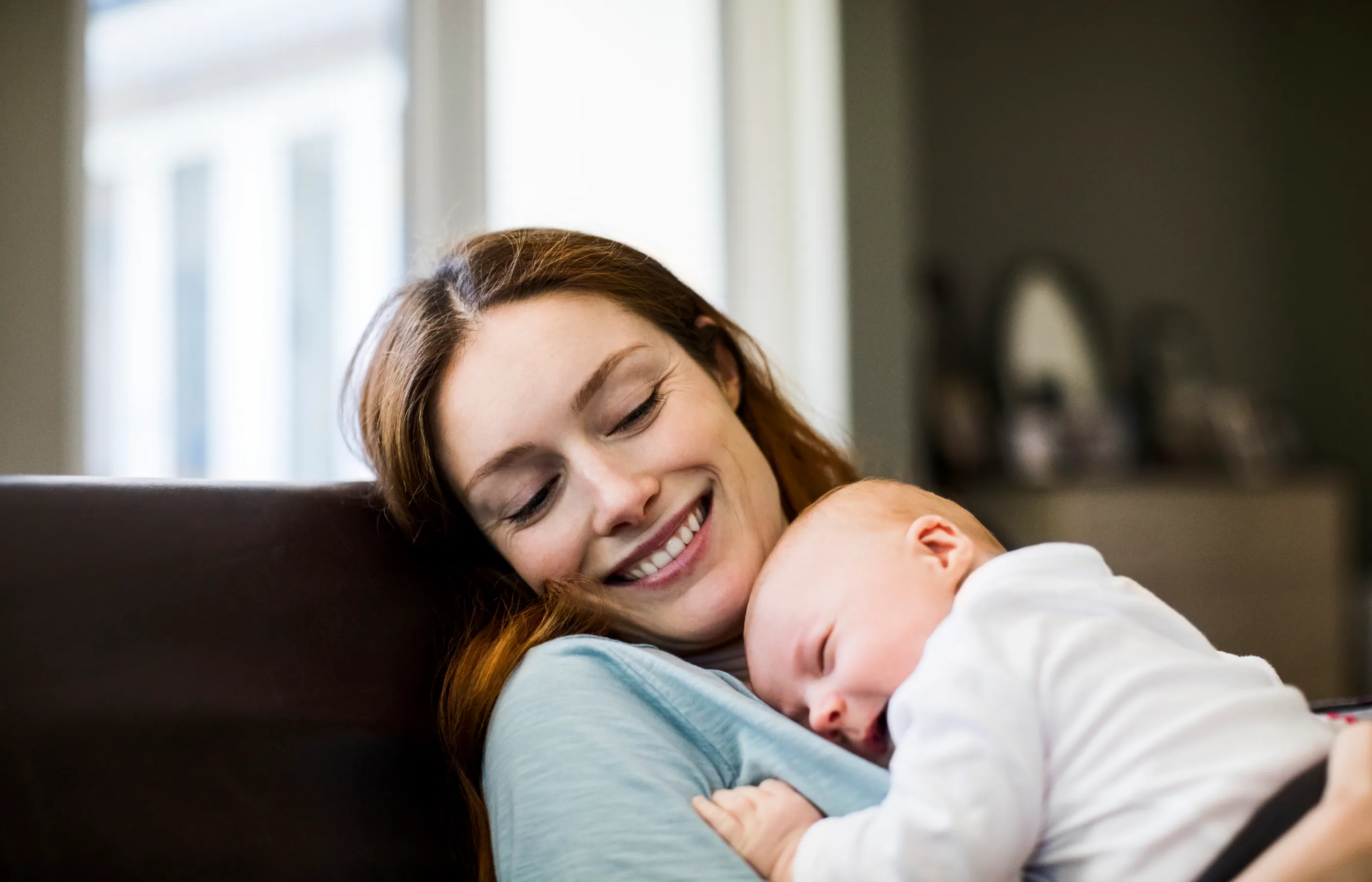 Young mother smiling and holding her newborn baby as she lies down on the couch. 