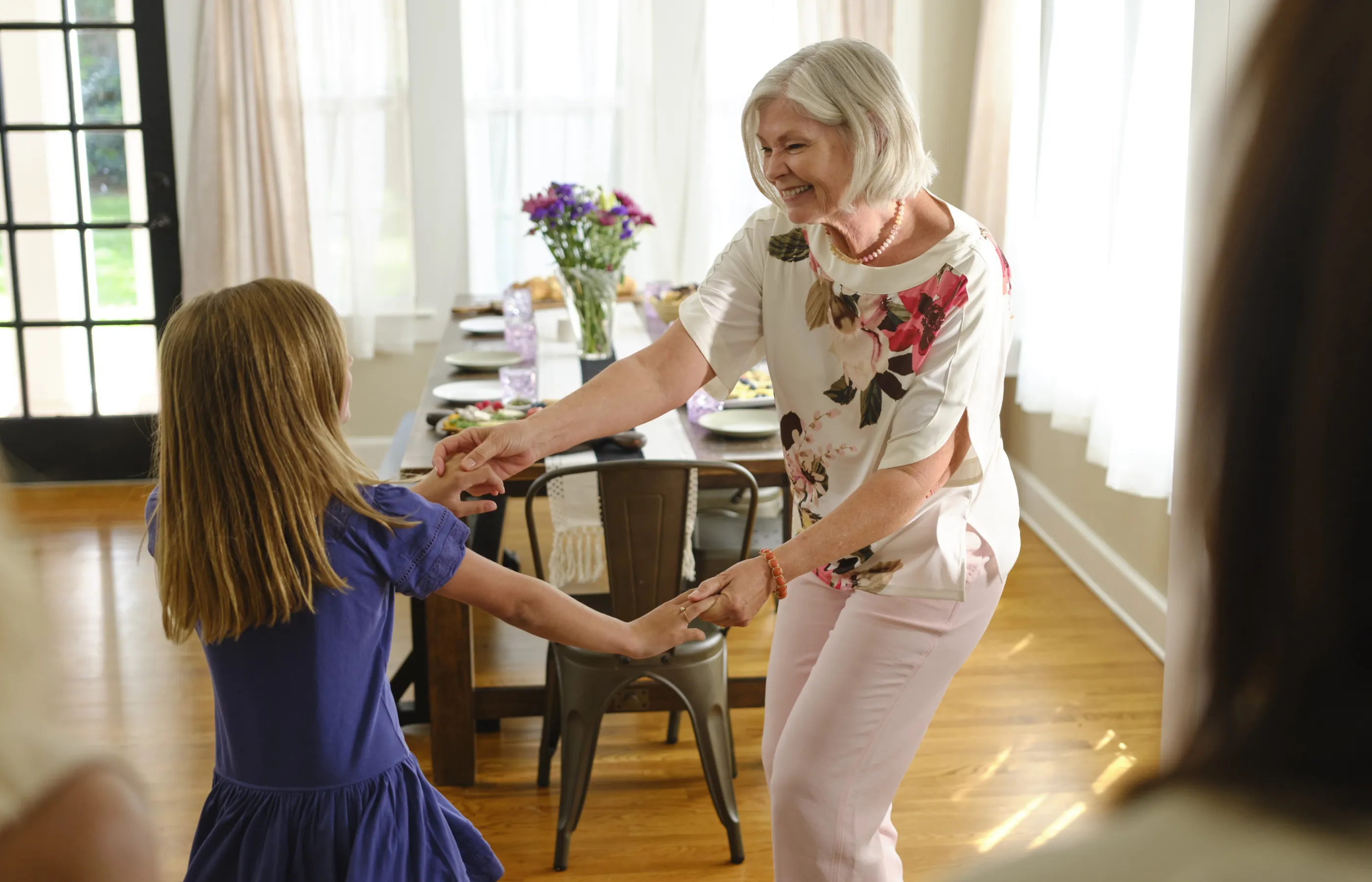 A senior woman dancing with her granddaughter in the dining room of a house.