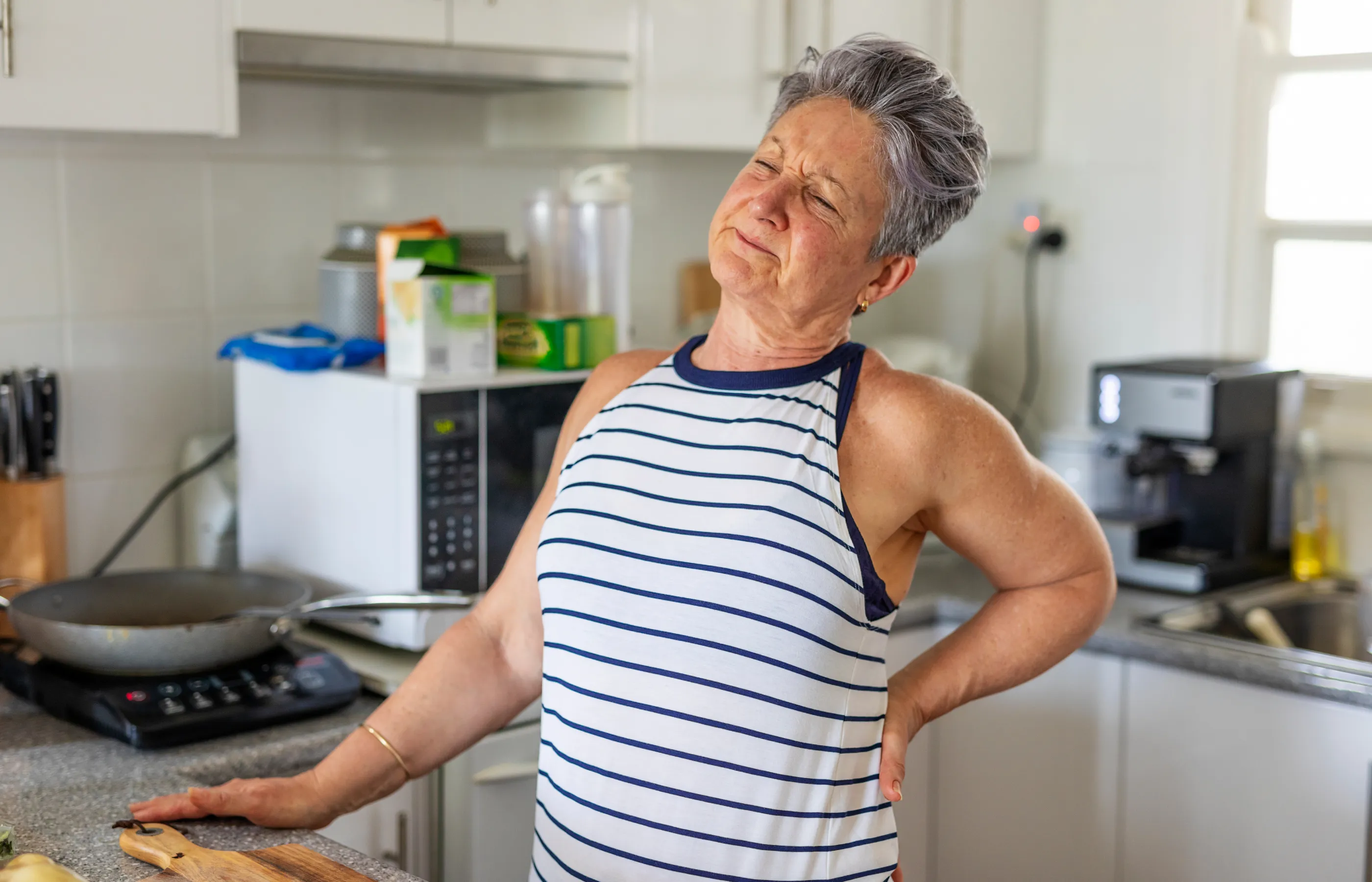 A senior woman is standing in her kitchen, preparing to cook, as she winces. She has one hand on the counter and the other on her lower back. 