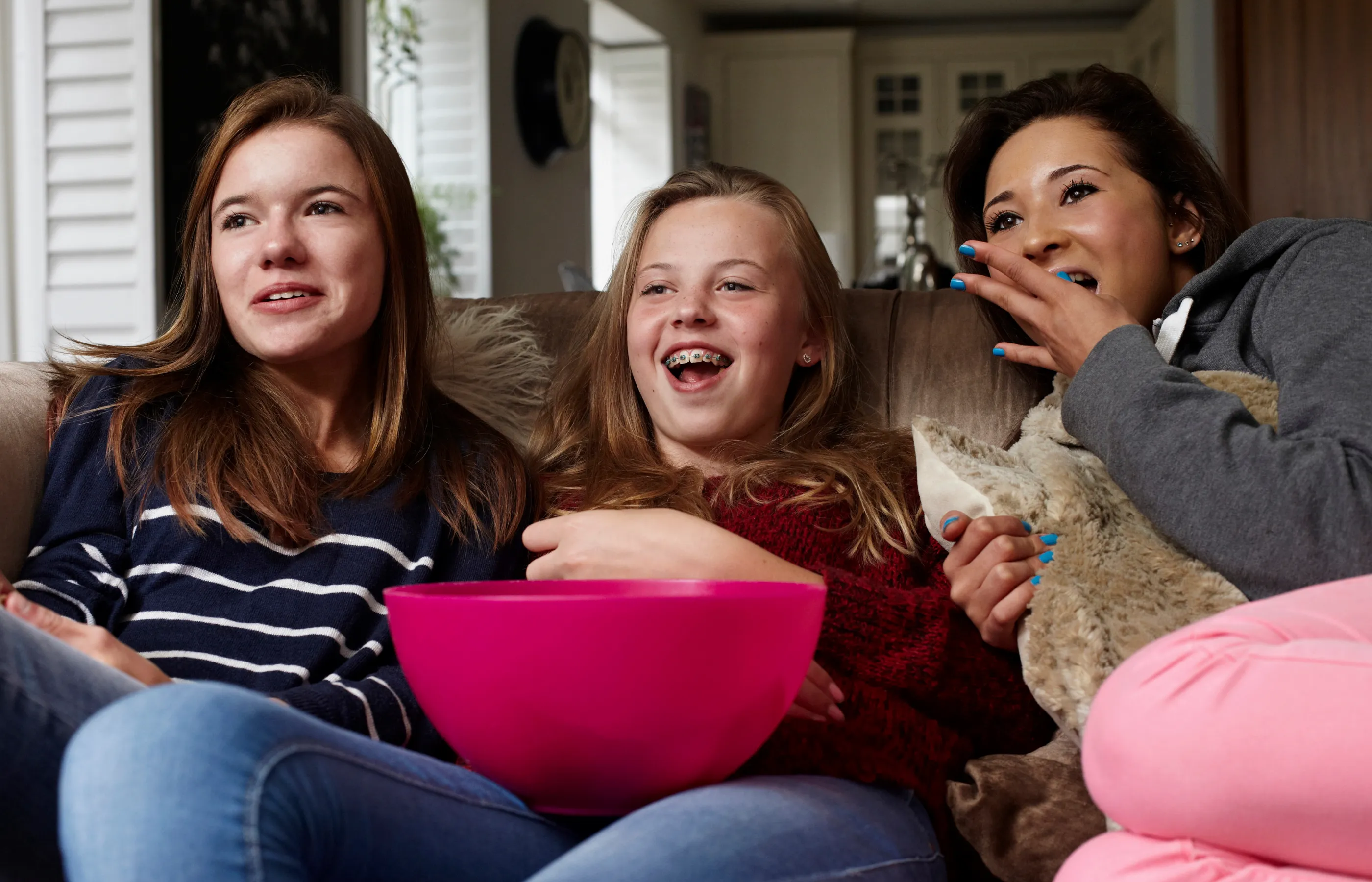 Three teenager girls are sitting on a couch eating popcorn as they watch television. 