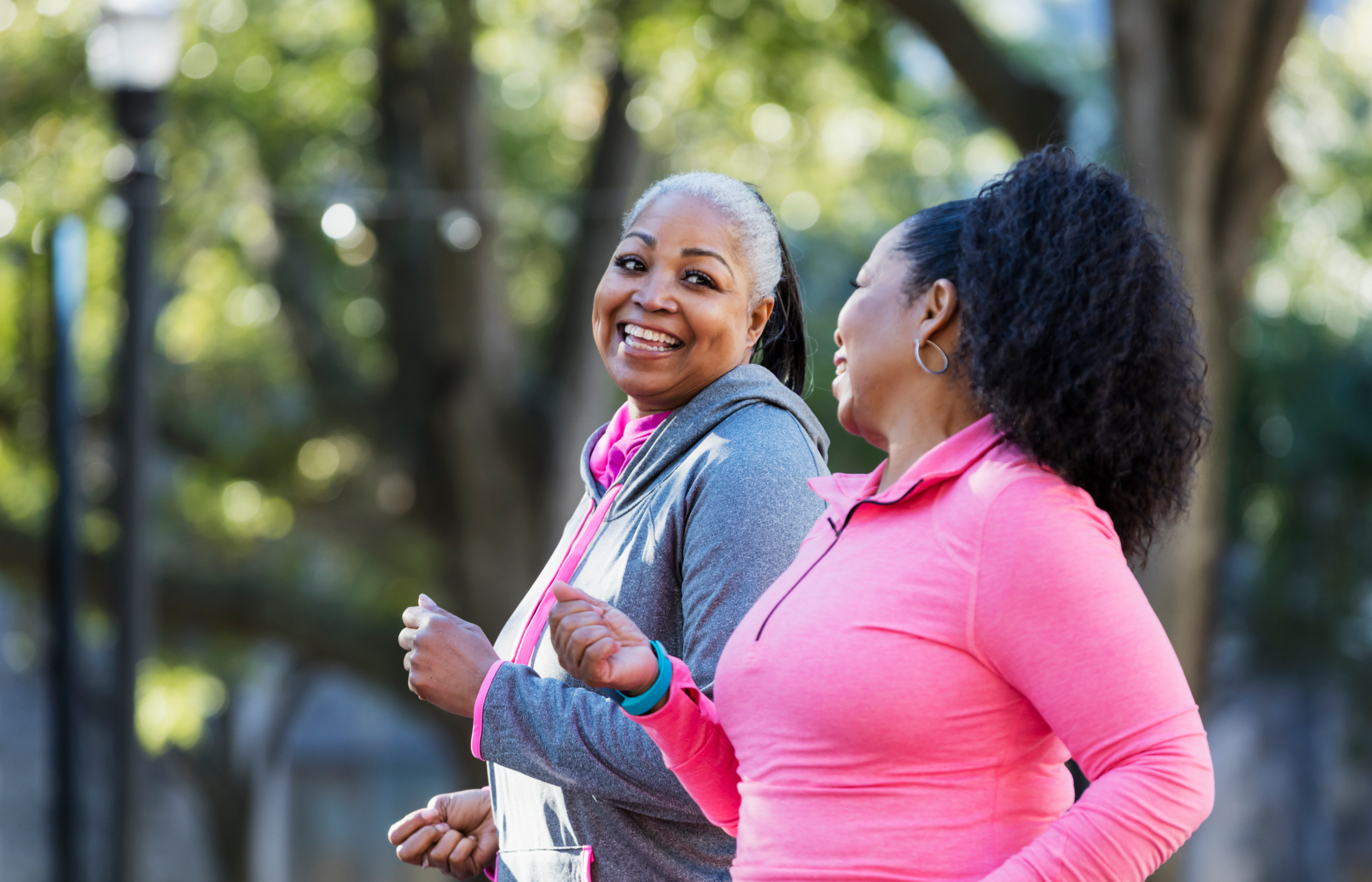 Two women, in pink workout attire, walking outdoors and smiling at each other. 
