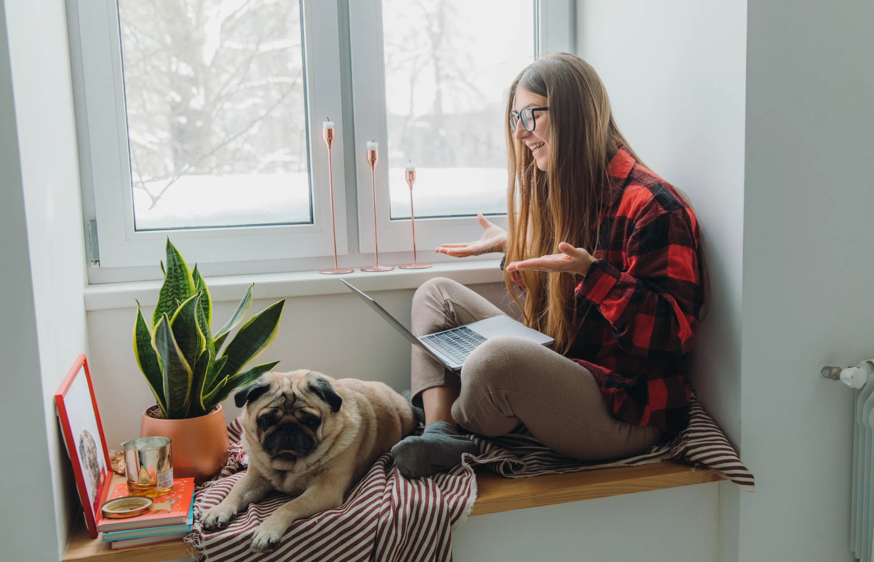 A woman sitting near a window wearing glasses while using a laptop.