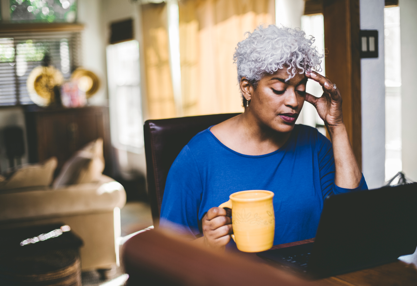 A woman sitting in front of a laptop with a mug rubs her head in pain.