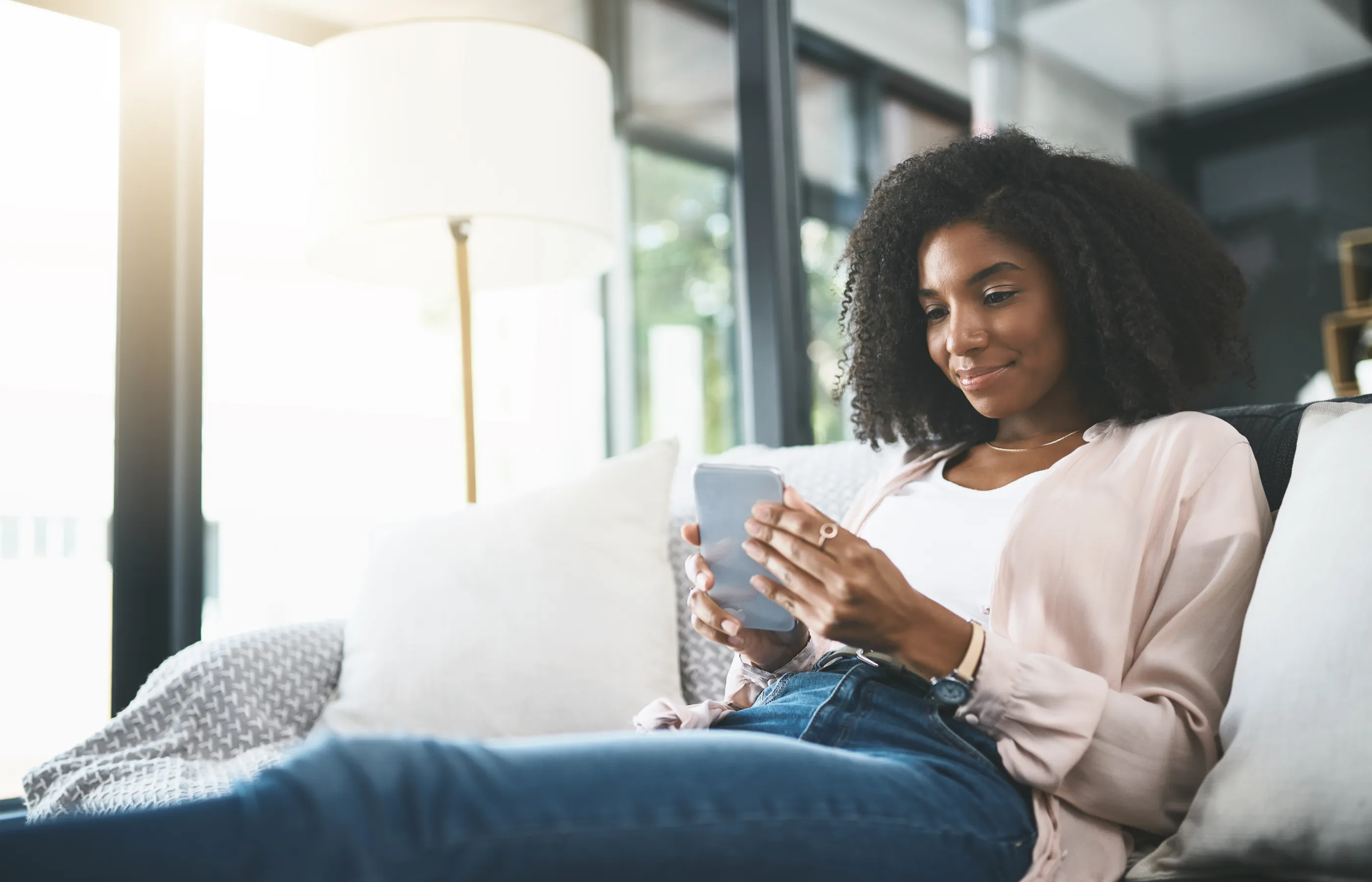 A woman smiles while looking at her phone near a window.