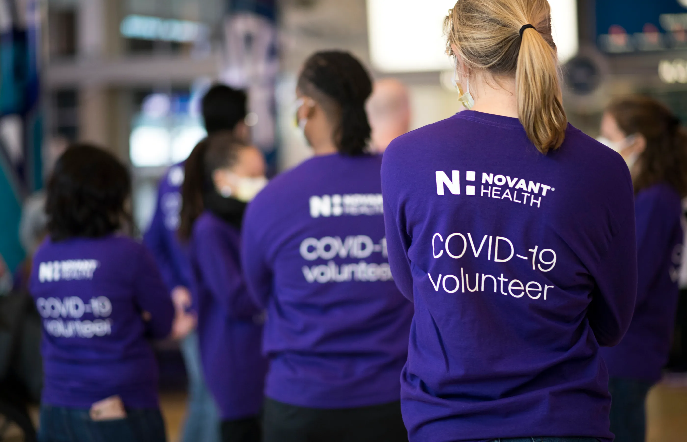 A group of volunteers are preparing for patients to arrive. They are all weaing masks and Novant Health Covid-19 volunteer t-shirts.  