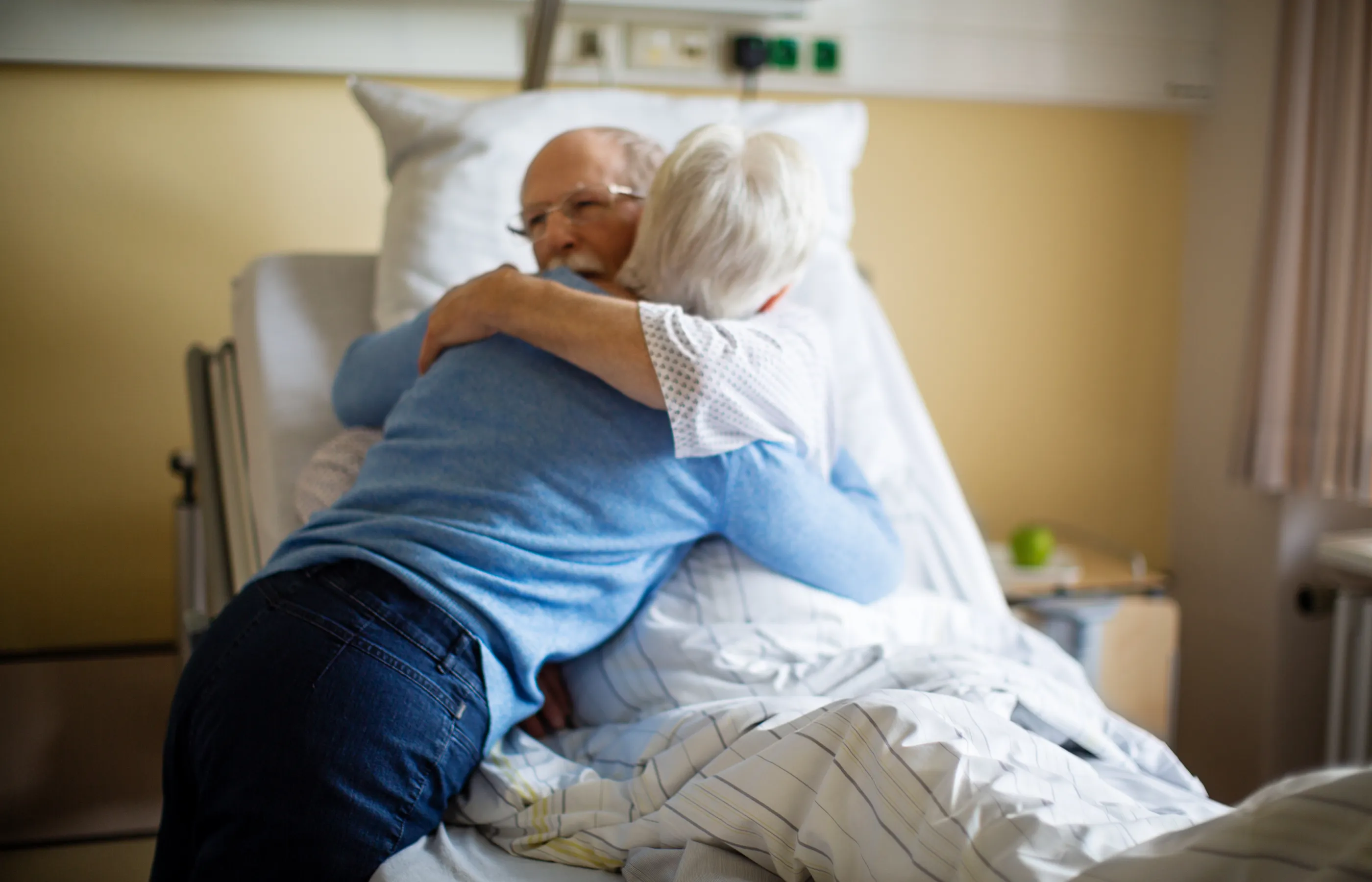 Patient and visitor in hospital room. The patient is lying in a hospital bed as the visitor hugs them. 