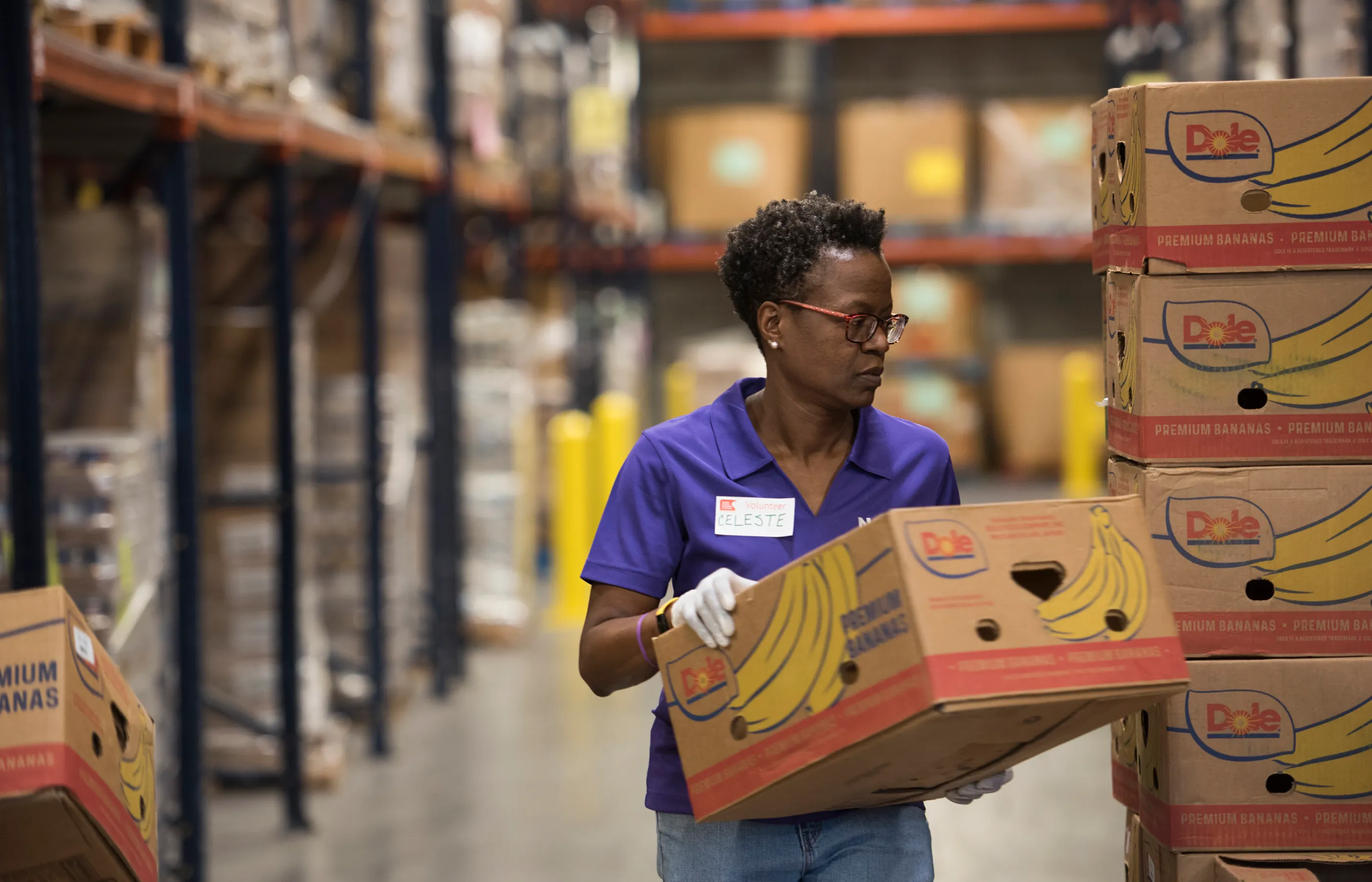 A woman is in a warehouse wearing a Novant Health shirt. She walks past a stack of boxes as she is carrying a Dole premium bananas box.