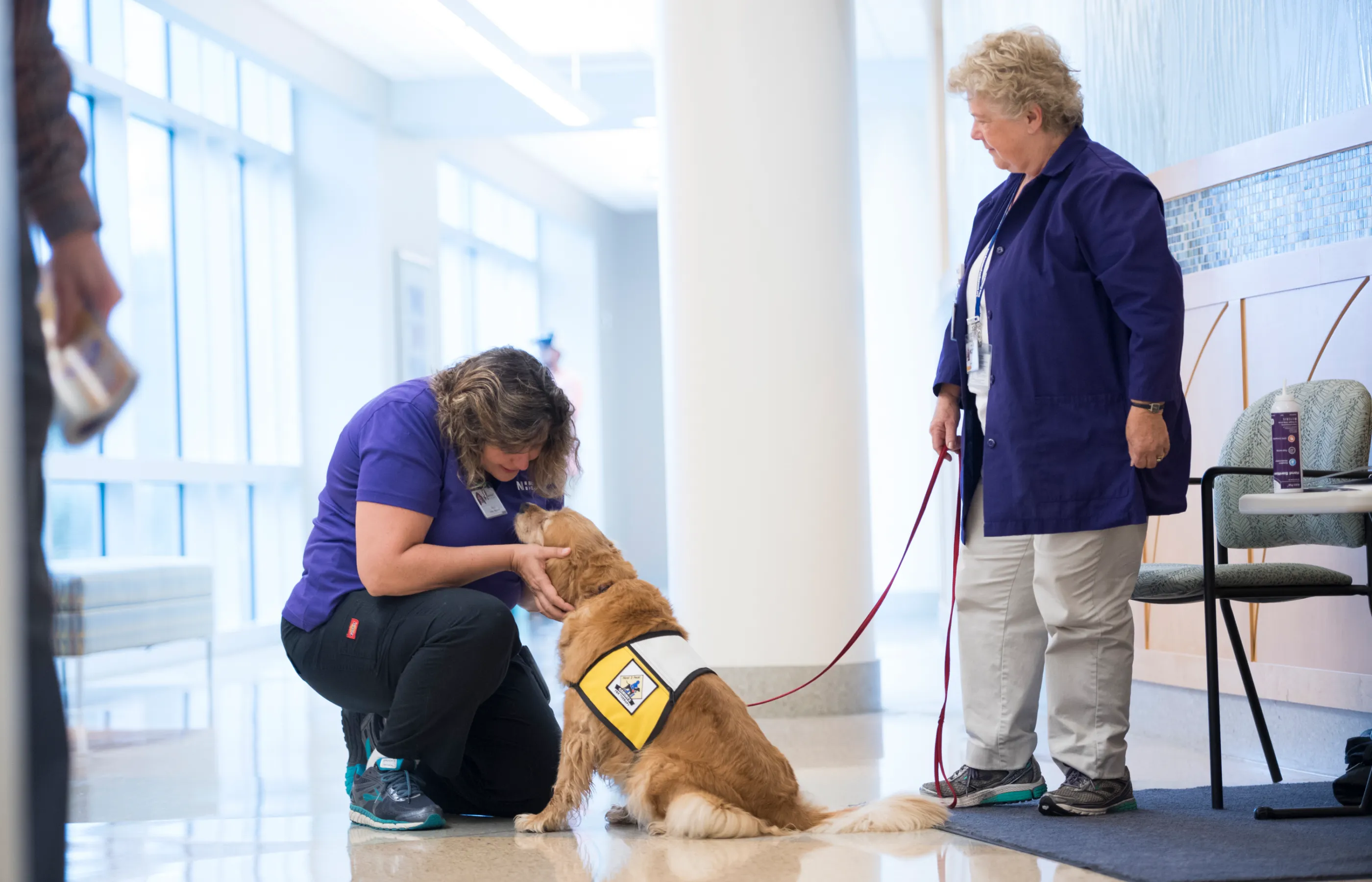 Two women are in the entry of a medical center. One is kneeling to greet a volunteer therapy dog, while the other is holding the dogs leash. 