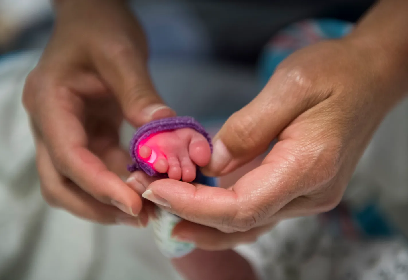 A health care provider has a newborns foot in their hand. The baby's foot has a red light senor on it that is wrapped in purple tape.
