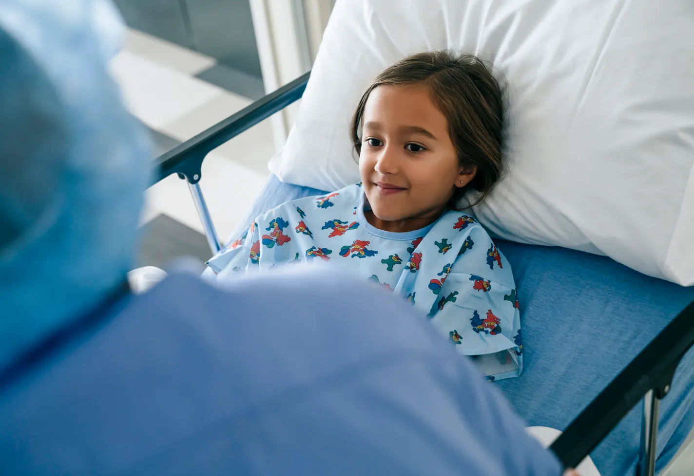  A young child is lying down in a hospital bed and looking up at a healthcare provider. 
